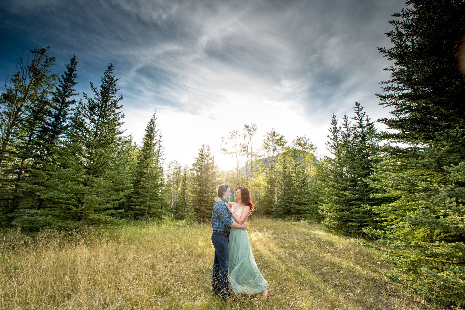 A couple embraces in a scenic mountain meadow surrounded by evergreen trees under dramatic cloudy skies in Banff, AB.
