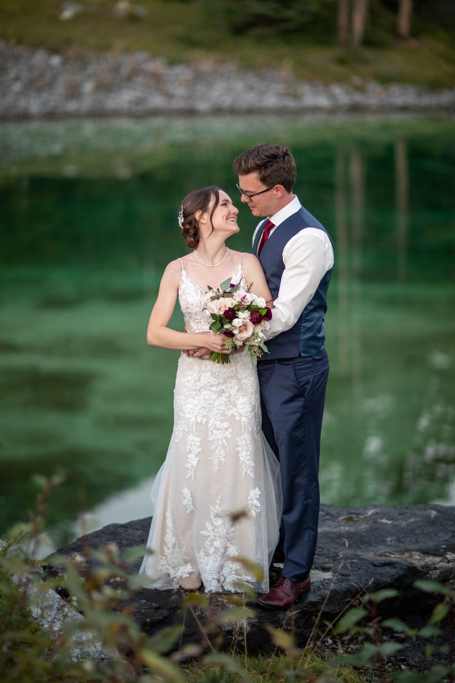 Newlywed couple shares romantic moments by a serene lake with emerald green waters and rocky shoreline at Silvertip Resort in Canmore, AB.