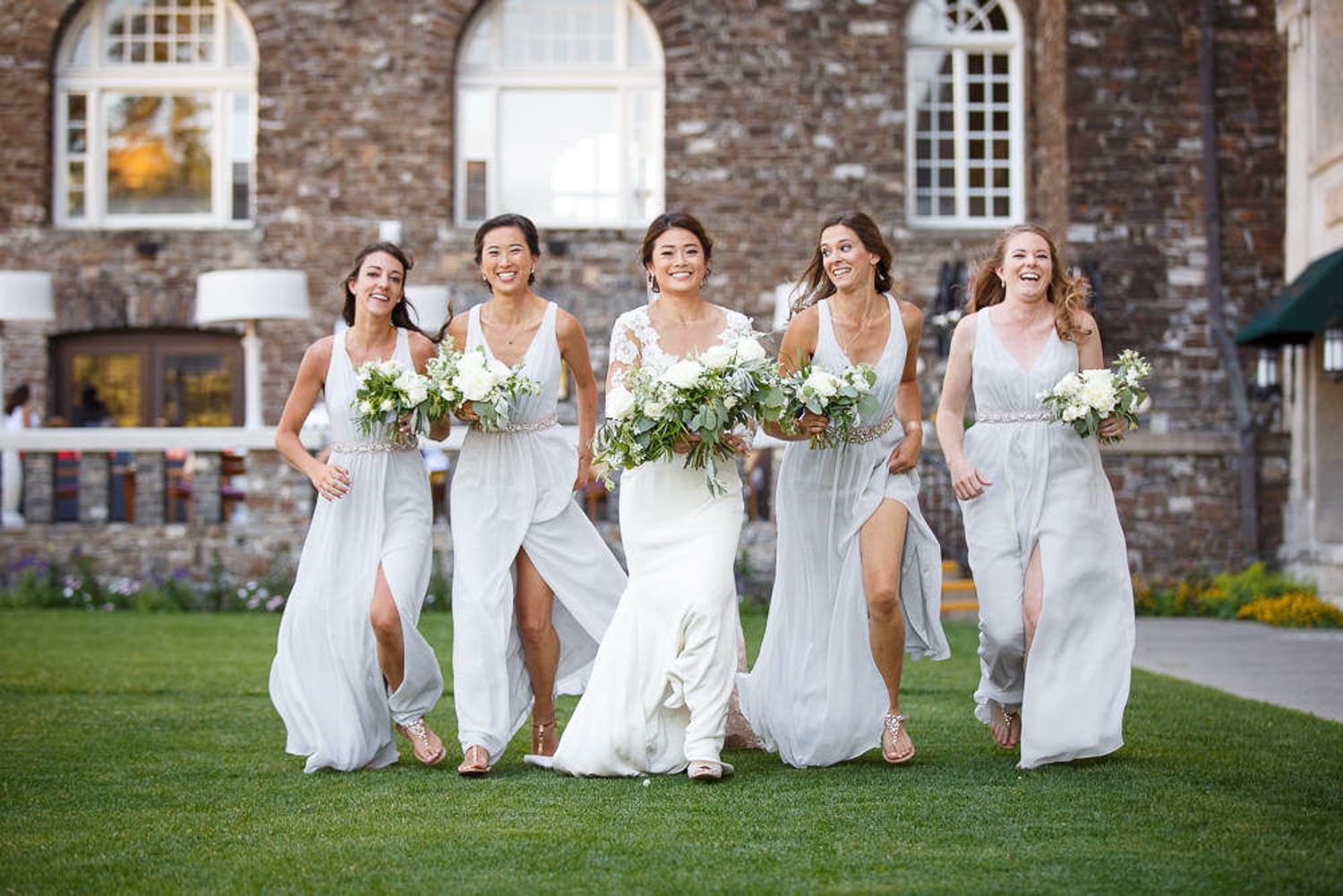 Bridesmaids in flowing white dresses run joyfully across a green lawn outside a stone building. Fairmont Banff springs hotel. Banff.