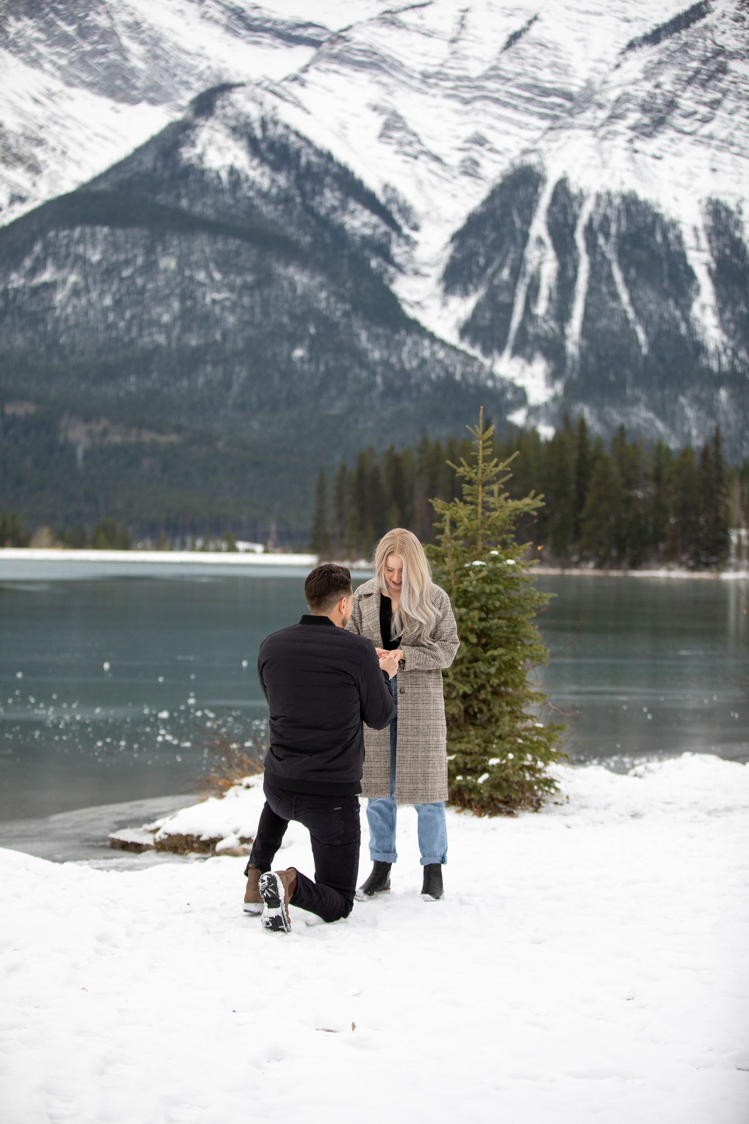 A winter proposal scene unfolds by a snow-covered lake with majestic mountains in the background in Canmore quarry lake.