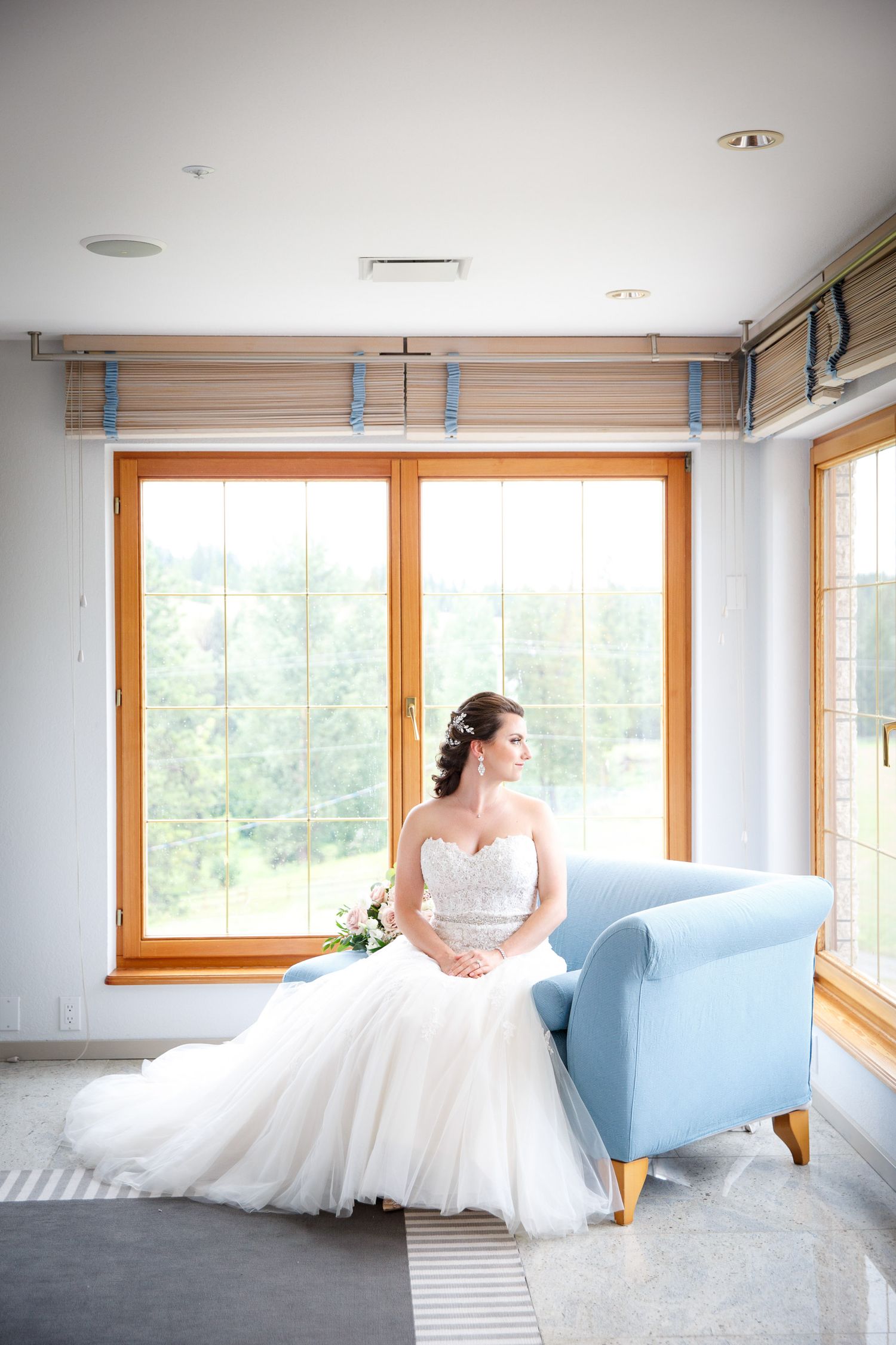 Elegant bride in a strapless white ball gown sitting gracefully on a light blue chair by large windows Cherry creek estate