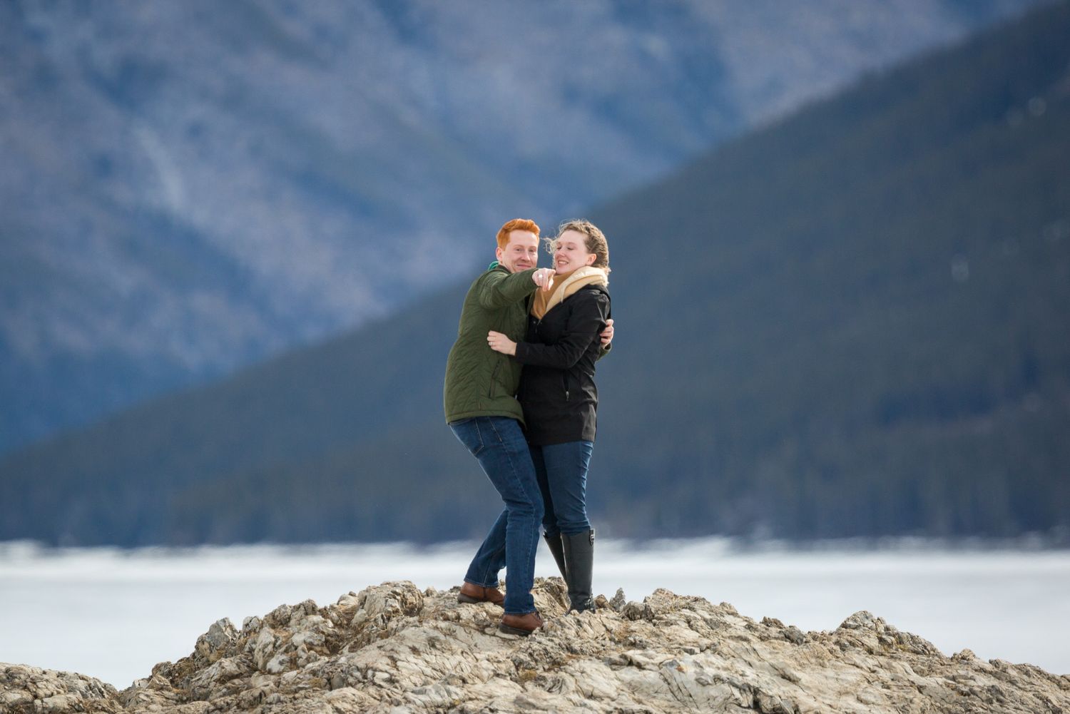 Couple embracing and smiling on rocky shore with mountain lake backdrop at Lake Minnewanka in Banff National Park.