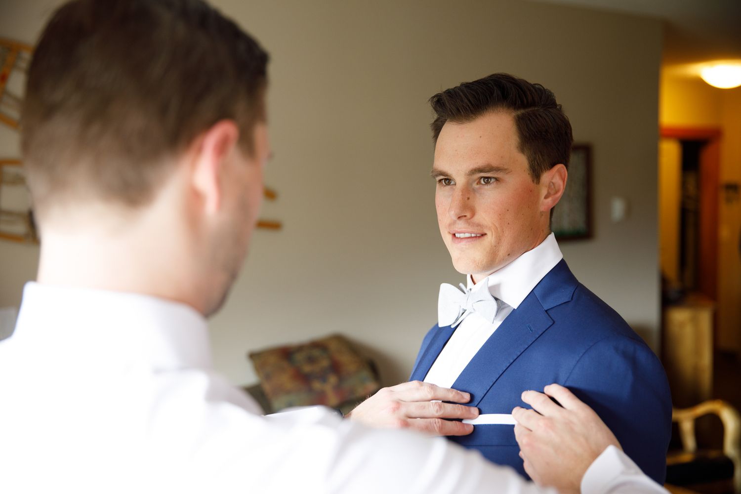 A groom in a blue suit gets help with adjusting his tie and boutonniere while getting ready for a wedding.