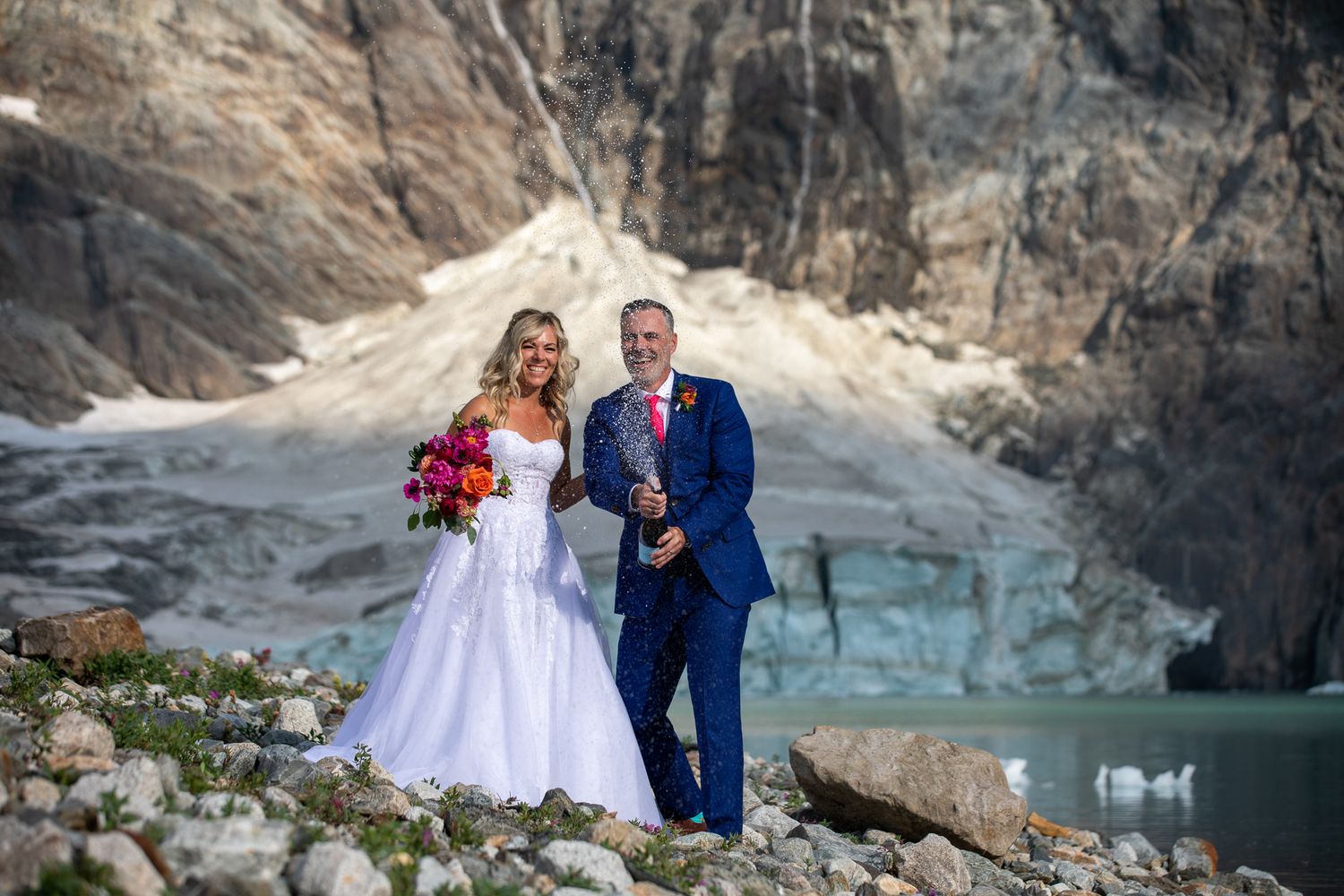 Bride and groom pose with champagne bottle before a glacier lake and rocky mountain backdrop on their wedding day at Lake-of-Hanging-Glacier