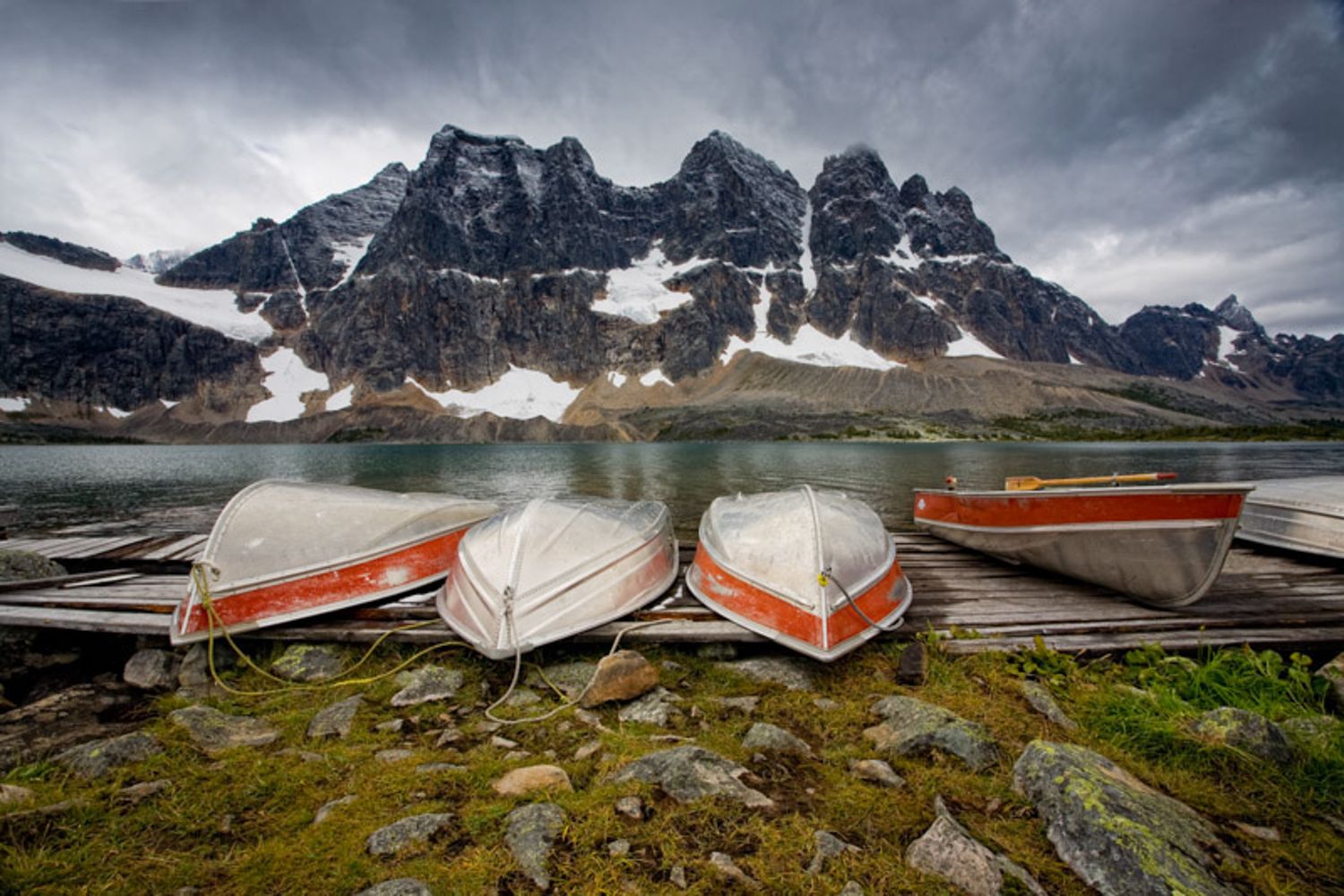 A panoramic view of rugged mountain peaks with snow patches and glacial lakes under dramatic cloudy skies. The ramparts in Jasper, AB.