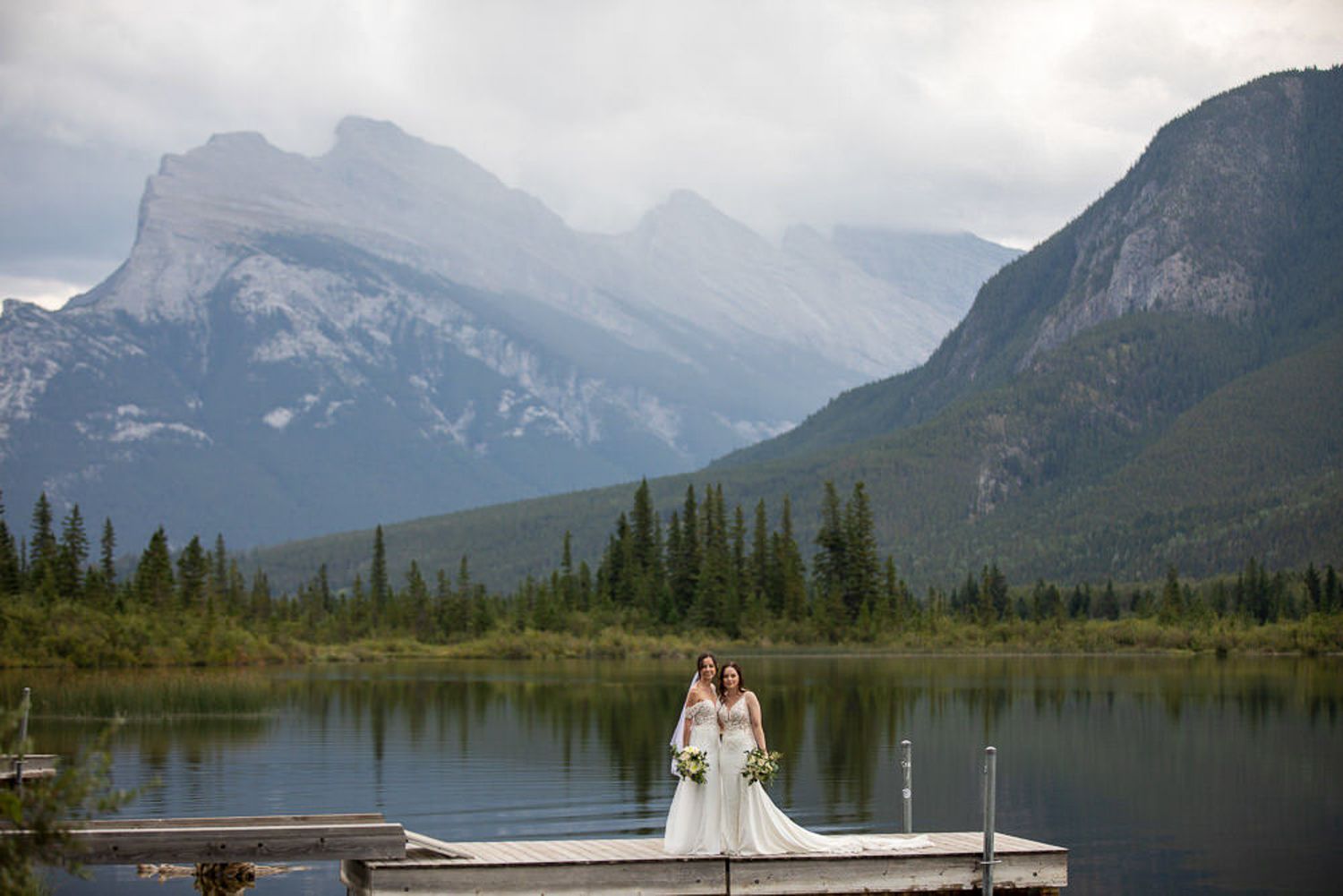 Two LGBTQ brides poses on a wooden dock surrounded by majestic mount Rundle and pristine Vermilion lake waters in Banff National Park.