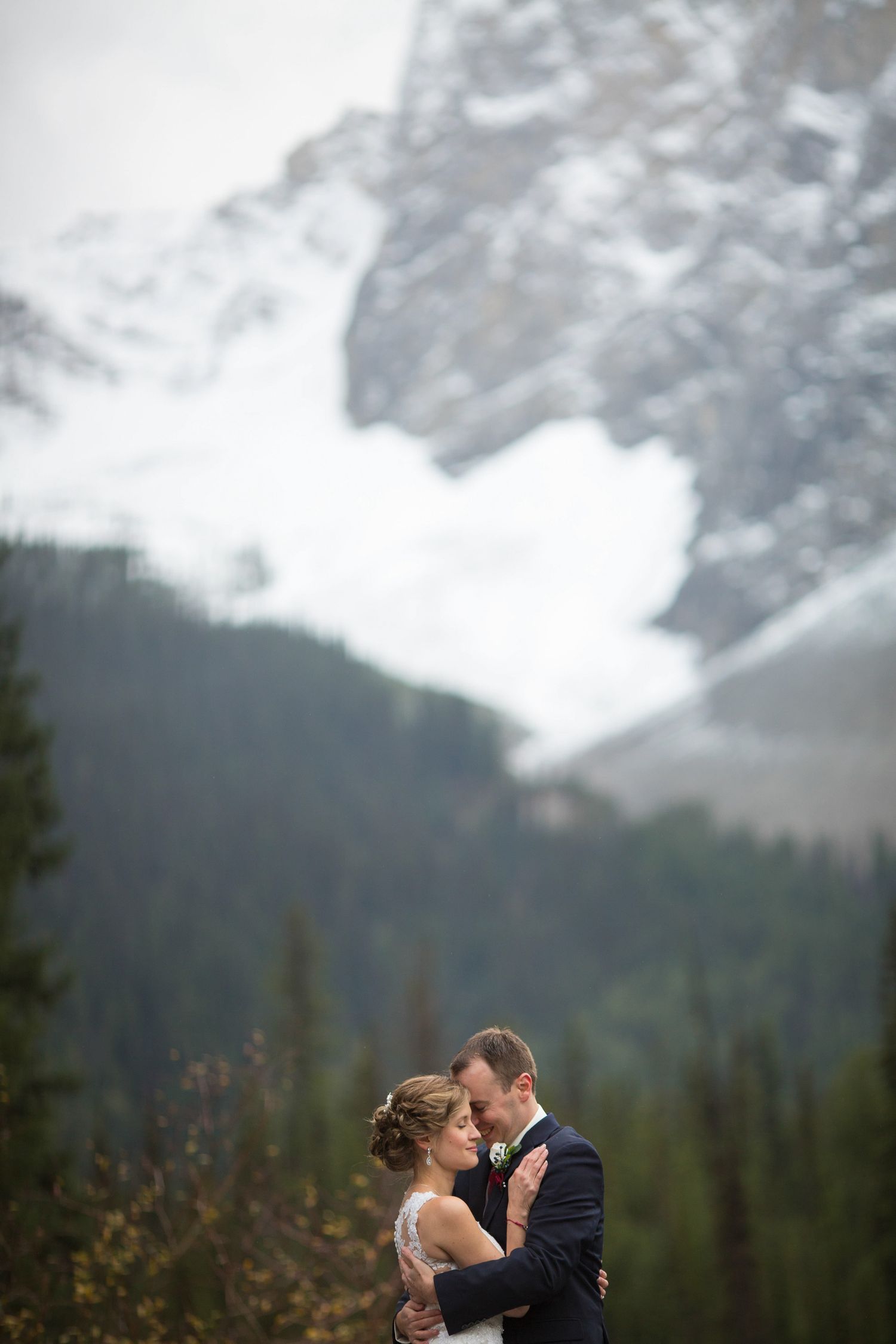 A romantic moment shared between a couple against a backdrop of misty mountain peaks and evergreen forest. Emerald Lake Lodge, Field BC