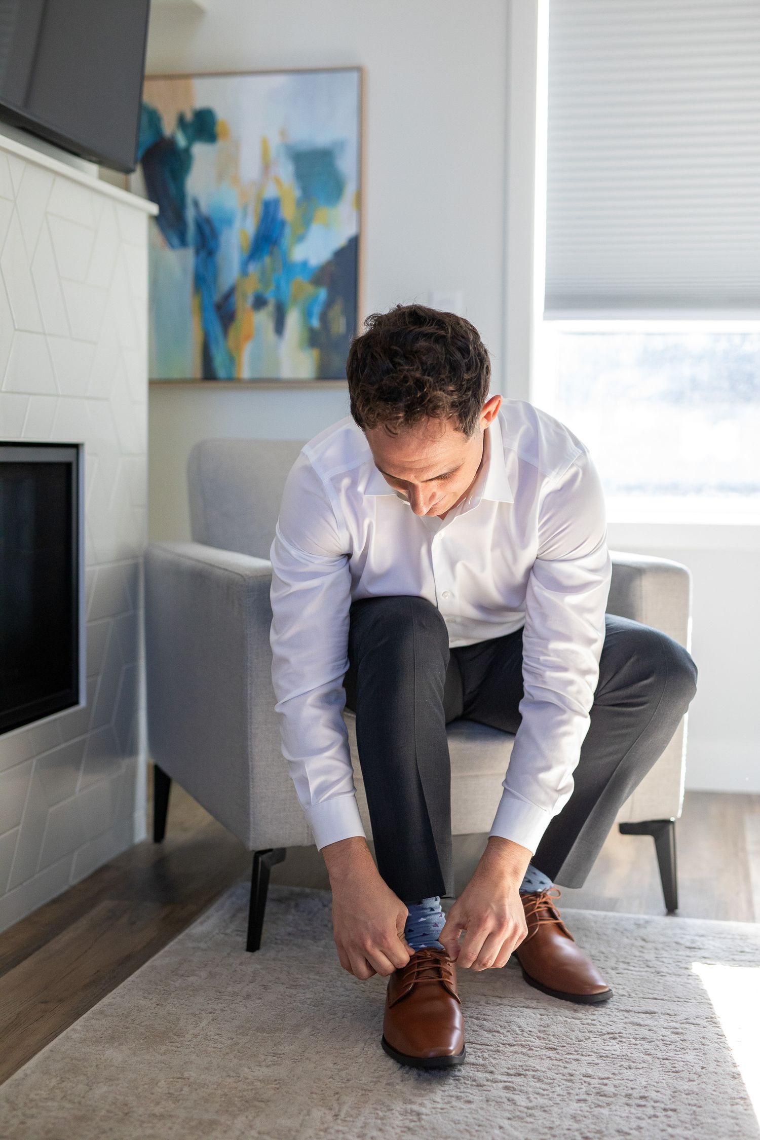 A groom in a white dress shirt and dark pants kneels on a light grey carpet while tying brown leather dress shoes. Canmore Solara resort.