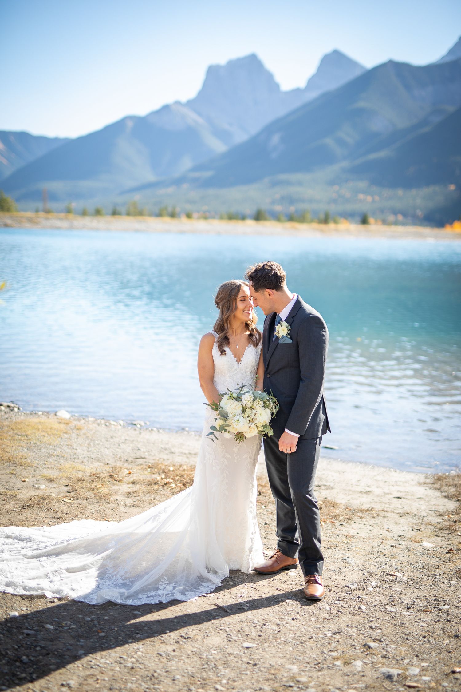 A couple shares a romantic moment on a scenic beach with majestic mountains and turquoise waters in the background at Quarry lake in Canmore