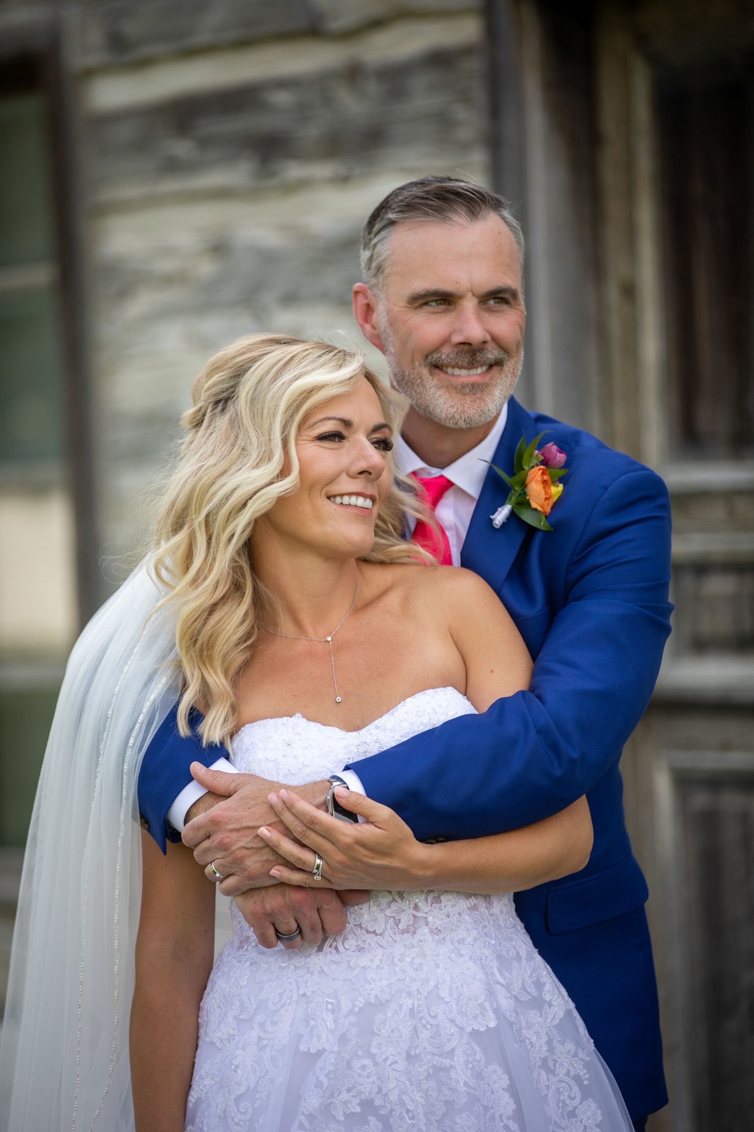 A smiling bride and groom embrace outdoors, she in a white lace gown with veil, he in a blue suit with pink tie. Eagle Ranch Resort, BC.