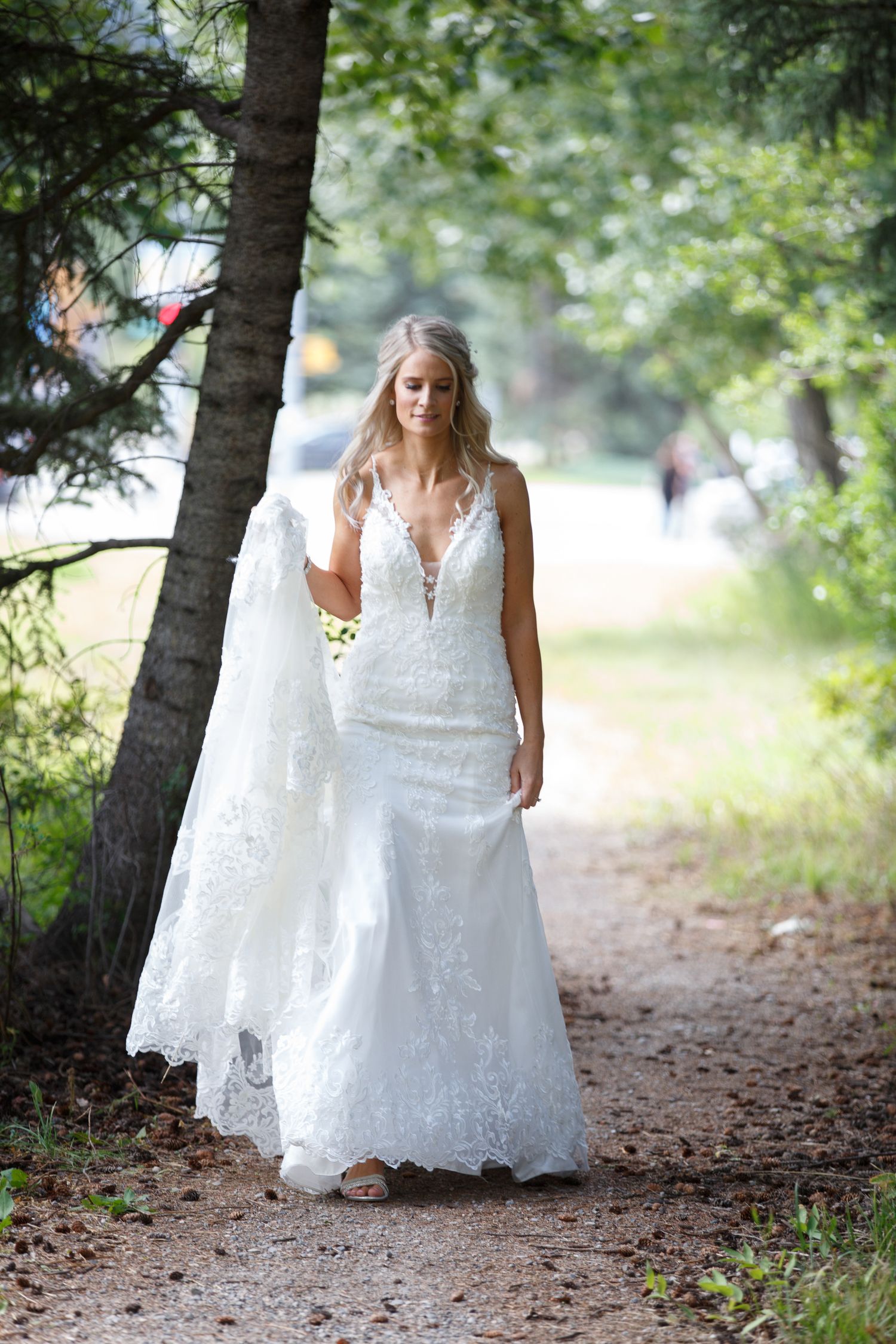 A bride in a flowing white lace wedding dress walks through a wooded path surrounded by green trees.