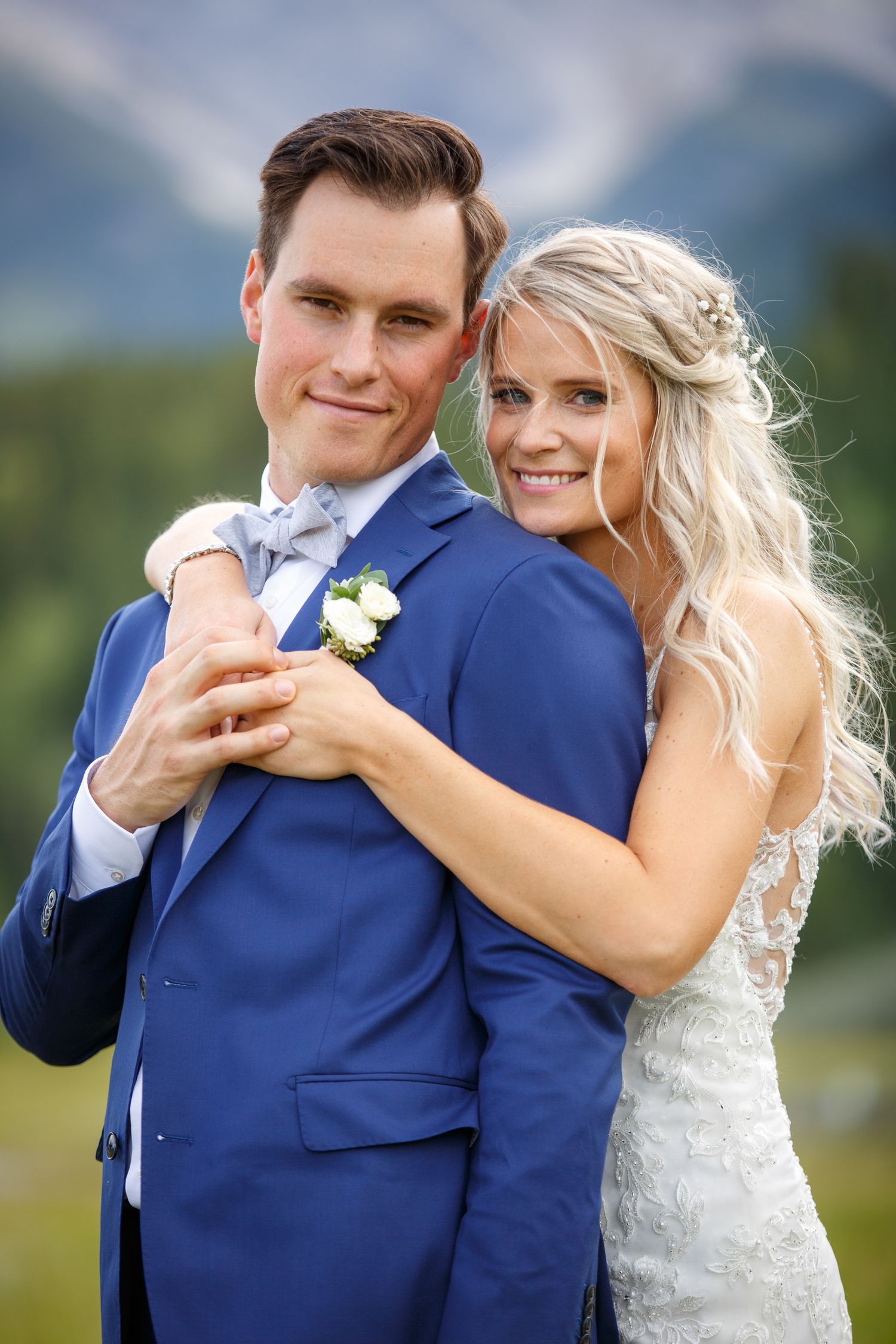 A romantic wedding portrait of a couple in a navy suit and white lace dress embracing outdoors with mountains in the background.
Canmore.