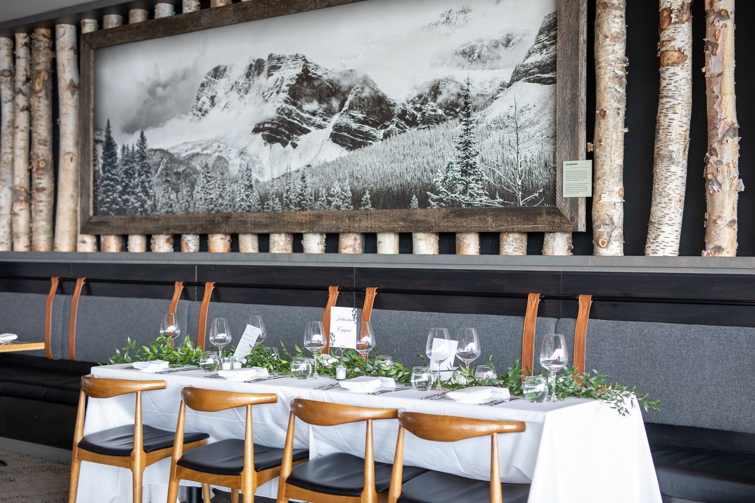Elegant restaurant dining table set with white linens and greenery beneath a black and white mountain landscape artwork.