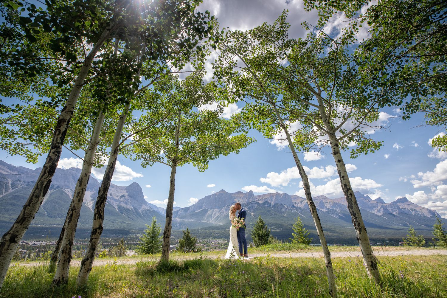 Couple embracing among tall aspen trees with stunning Rocky Mountain peaks and blue sky backdrop in Canmore, Alberta.