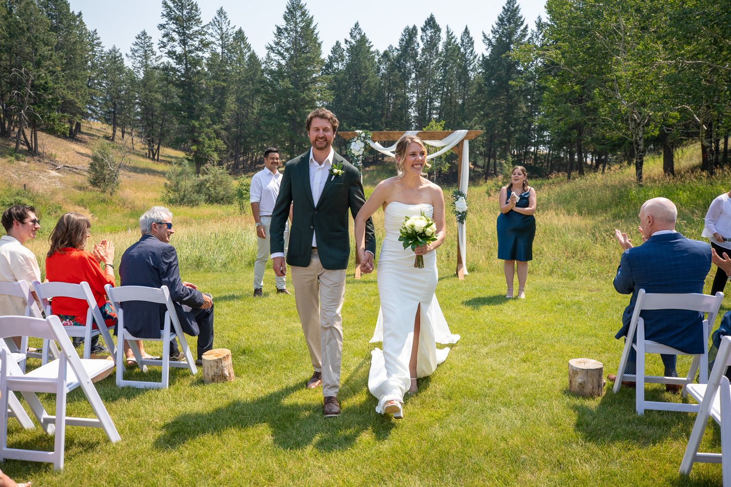 Bride and groom walk down outdoor aisle at sunny mountain wedding ceremony with seated guests on white chairs at Swansea Ranch in Invermere
