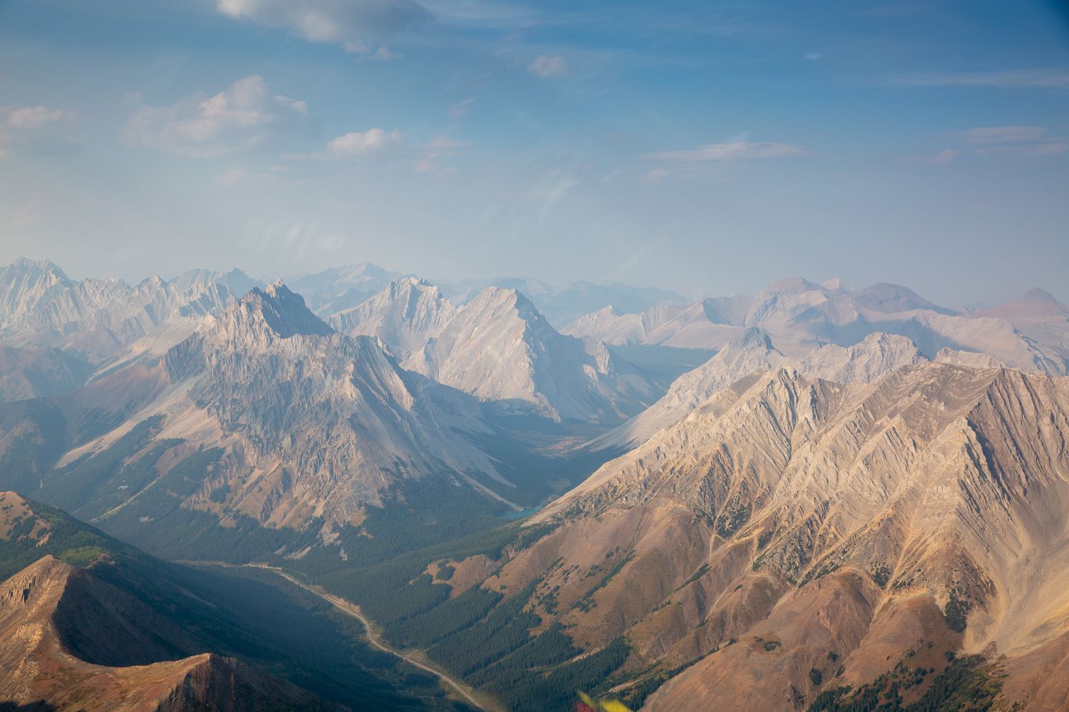 Aerial view of rugged mountain peaks and valleys under a hazy blue sky in a vast wilderness landscape in Kananaskis, Alberta.
