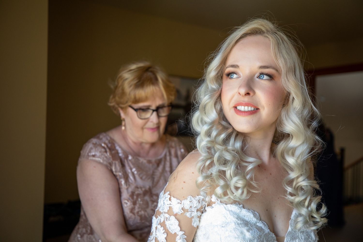 Bride with curly blonde hair in lace gown being helped by older woman in pink dress during wedding preparations In Canmore
