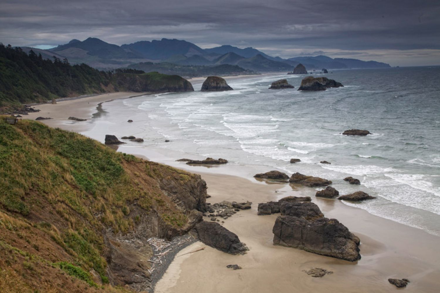 Dramatic aerial view of rugged Oregon coastline with rocky outcrops, sandy beaches and misty mountains in distance.