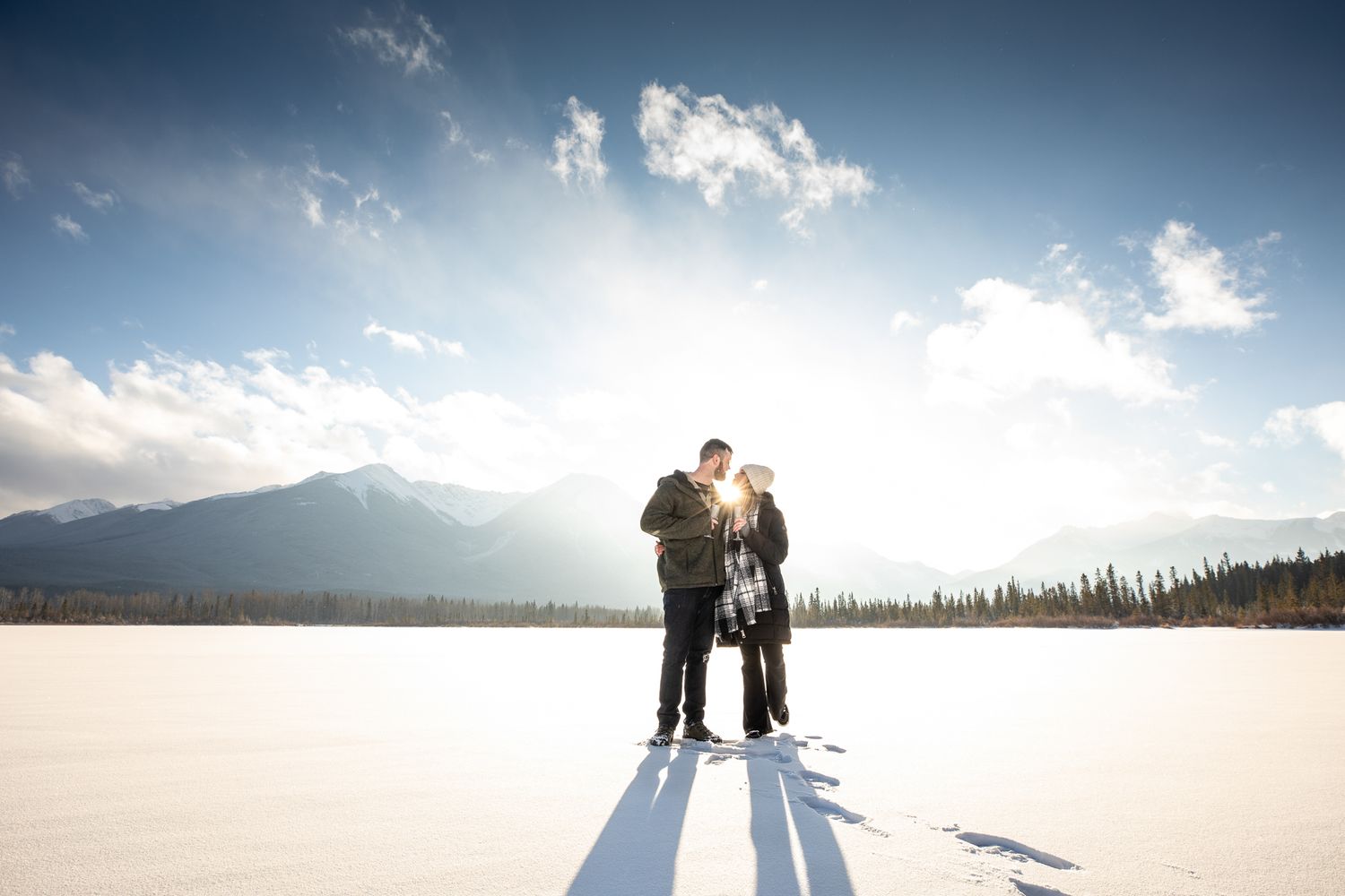 A couple stands together casting long shadows on a snowy mountain landscape at sunset at Vermilion Lakes in Banff, AB.