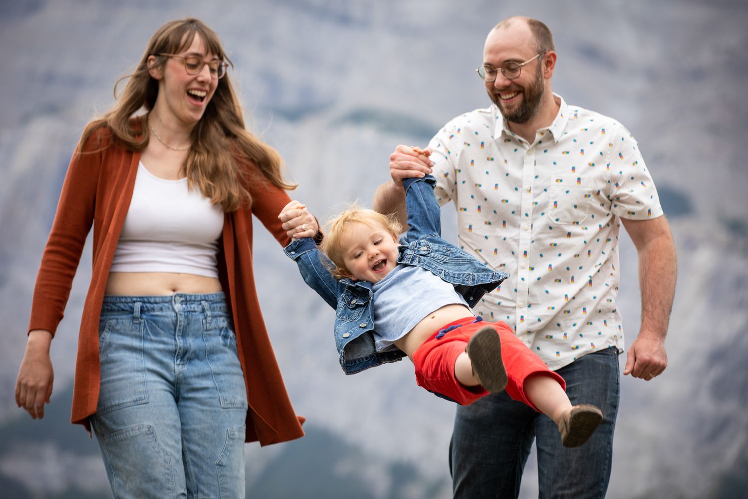 A joyful family in casual clothing holds hands and dances together outdoors against a cloudy sky background in Banff, AB.