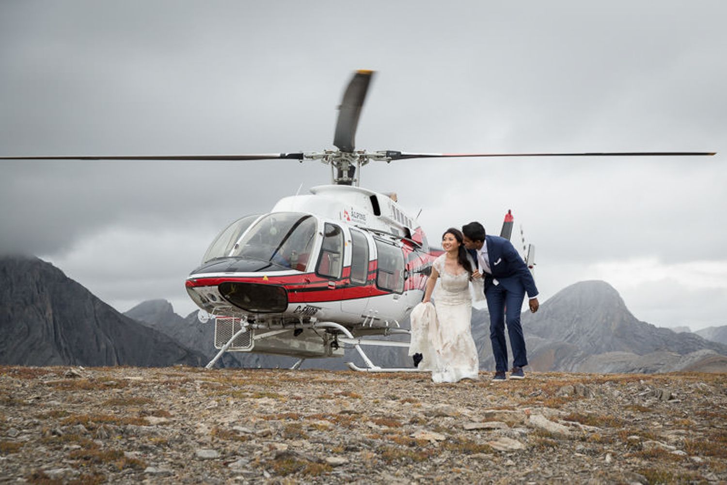A white and red helicopter landed on a rocky mountain peak with wedding couple standing beside Grotto Mountain Canmore, AB