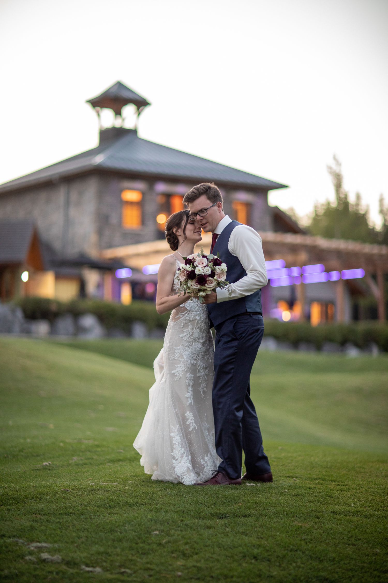 A romantic moment between a couple on a grassy hill at dusk with the beautiful Silvertip venue in the background in Canmore, AB.