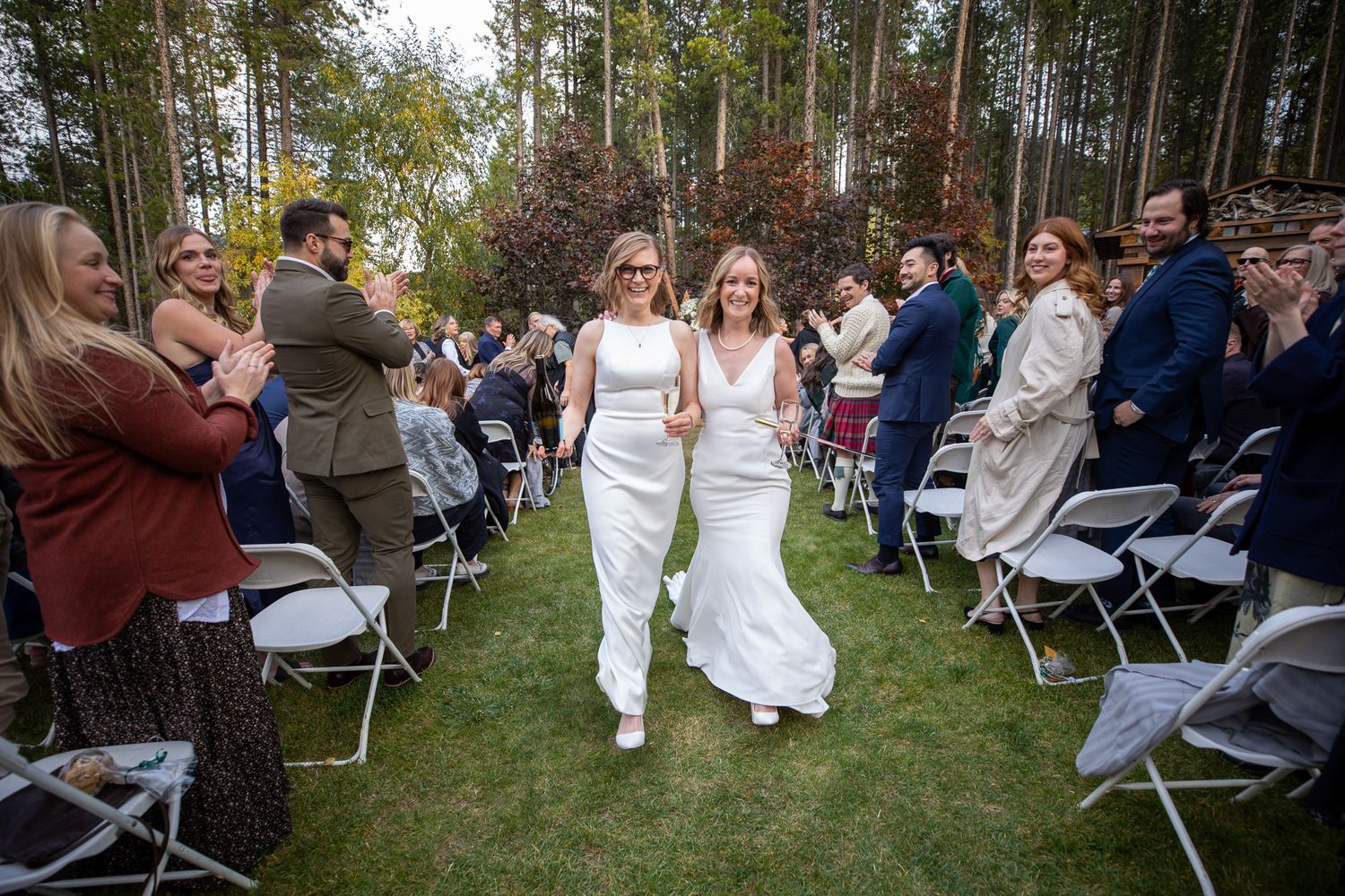 Wedding ceremony guests seated in folding chairs outdoors among tall pine trees at a forest venue. Earl Grey Lodge Panorama BC