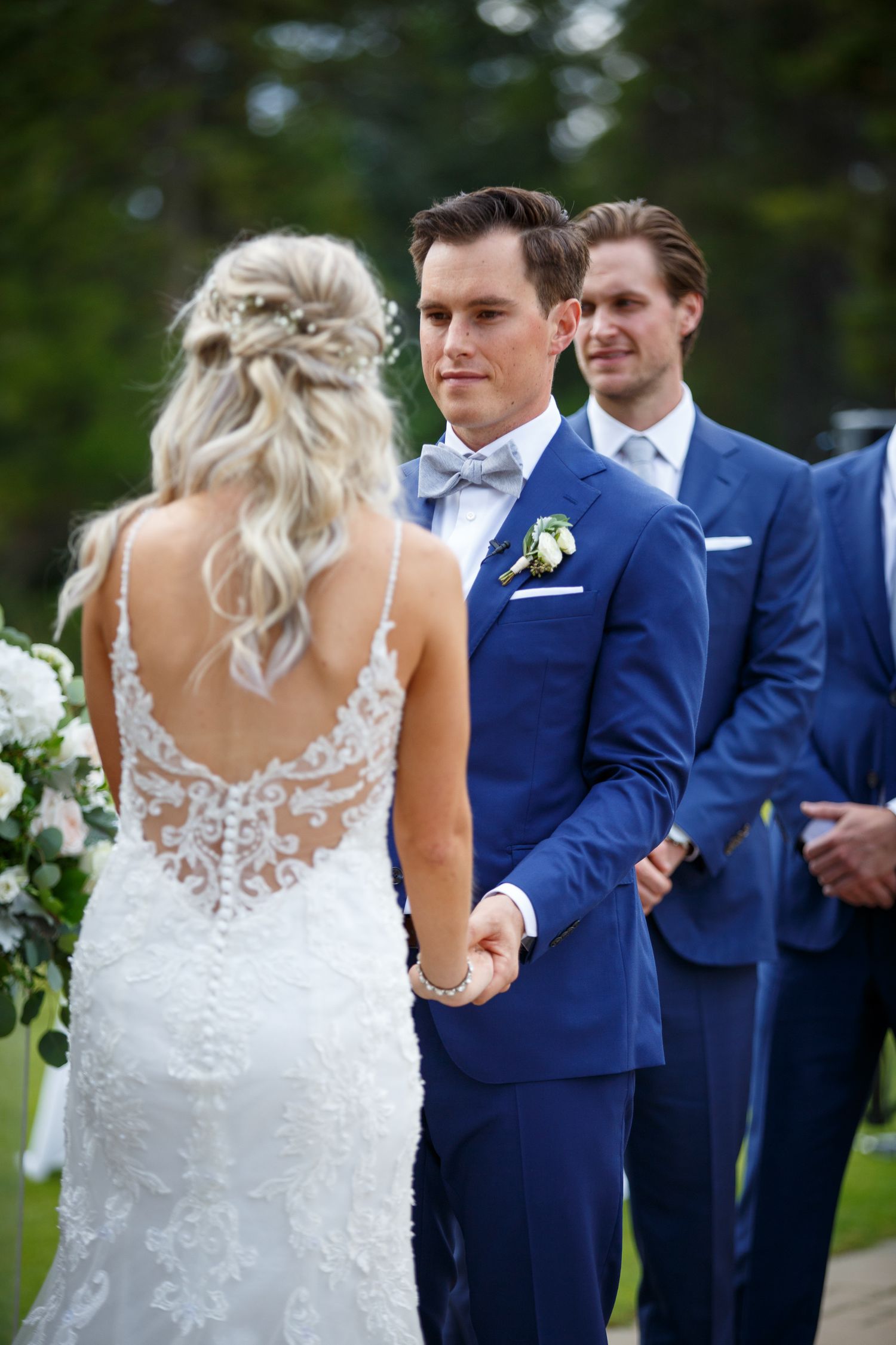 Wedding party members in navy suits and a white lace dress standing together at an outdoor ceremony.