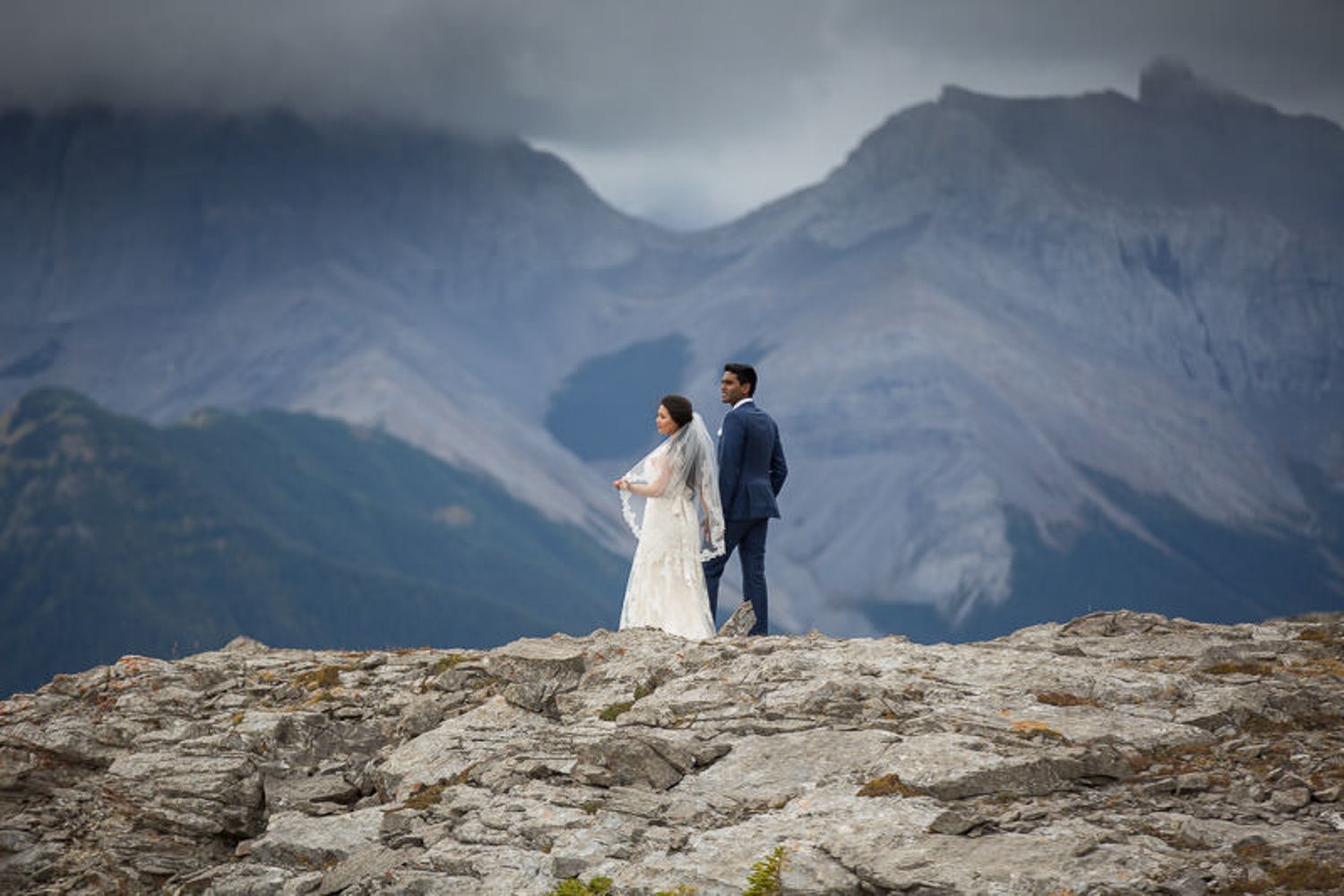 A couple stands on a rocky mountain ledge overlooking a dramatic mountain landscape in Banff National Park.