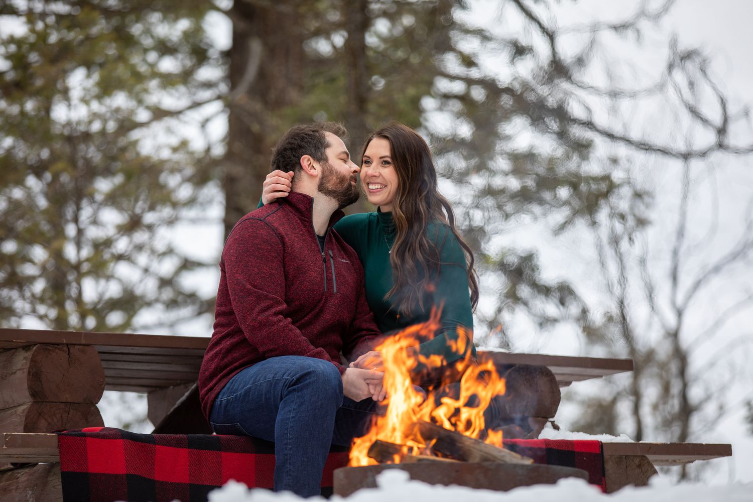 A couple sits on a wooden bench enjoying hot drinks by a cozy campfire on a winter day in the forest at Grey Wolf Panorama, BC