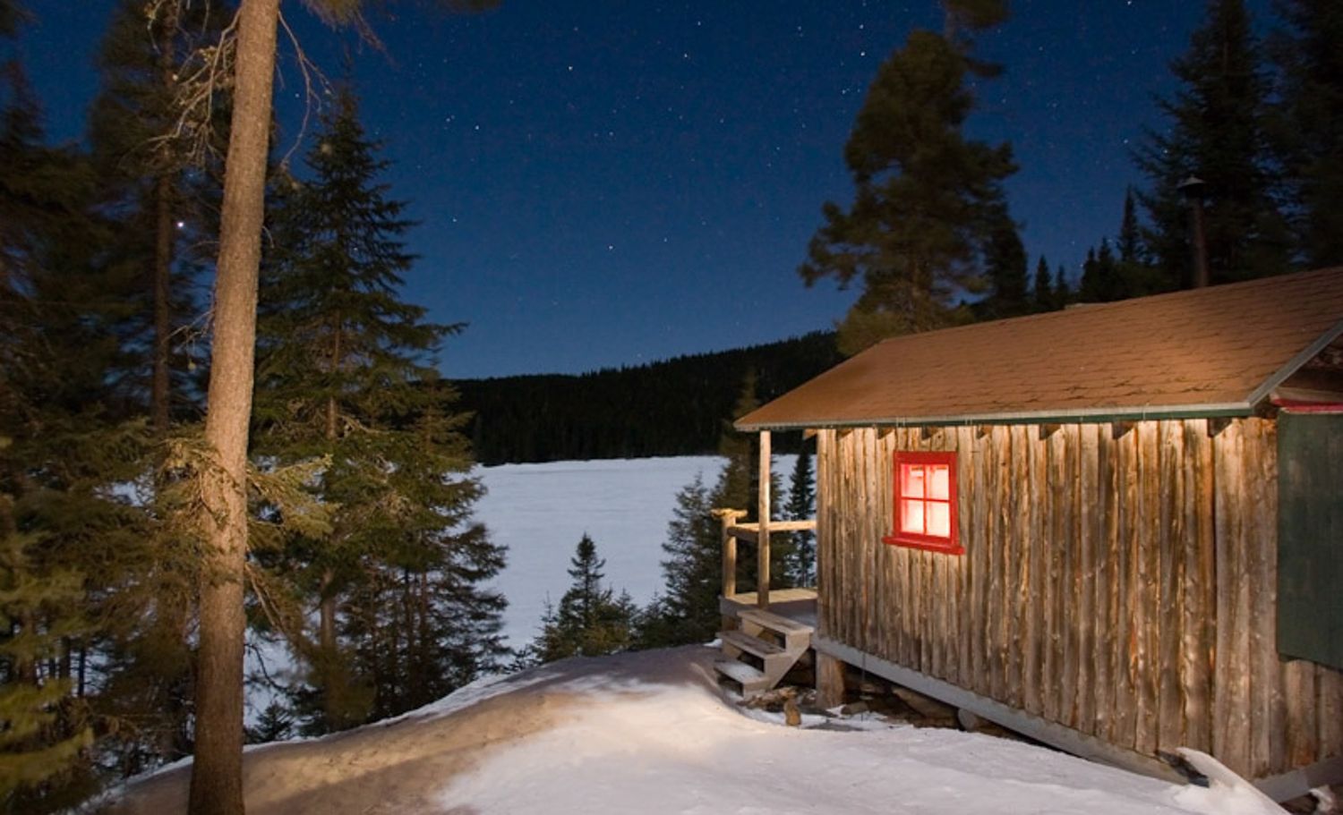 A rustic wooden cabin sits nestled among tall pine trees in a snowy winter landscape at dusk in Quebec countryside, Charlevoix, QC