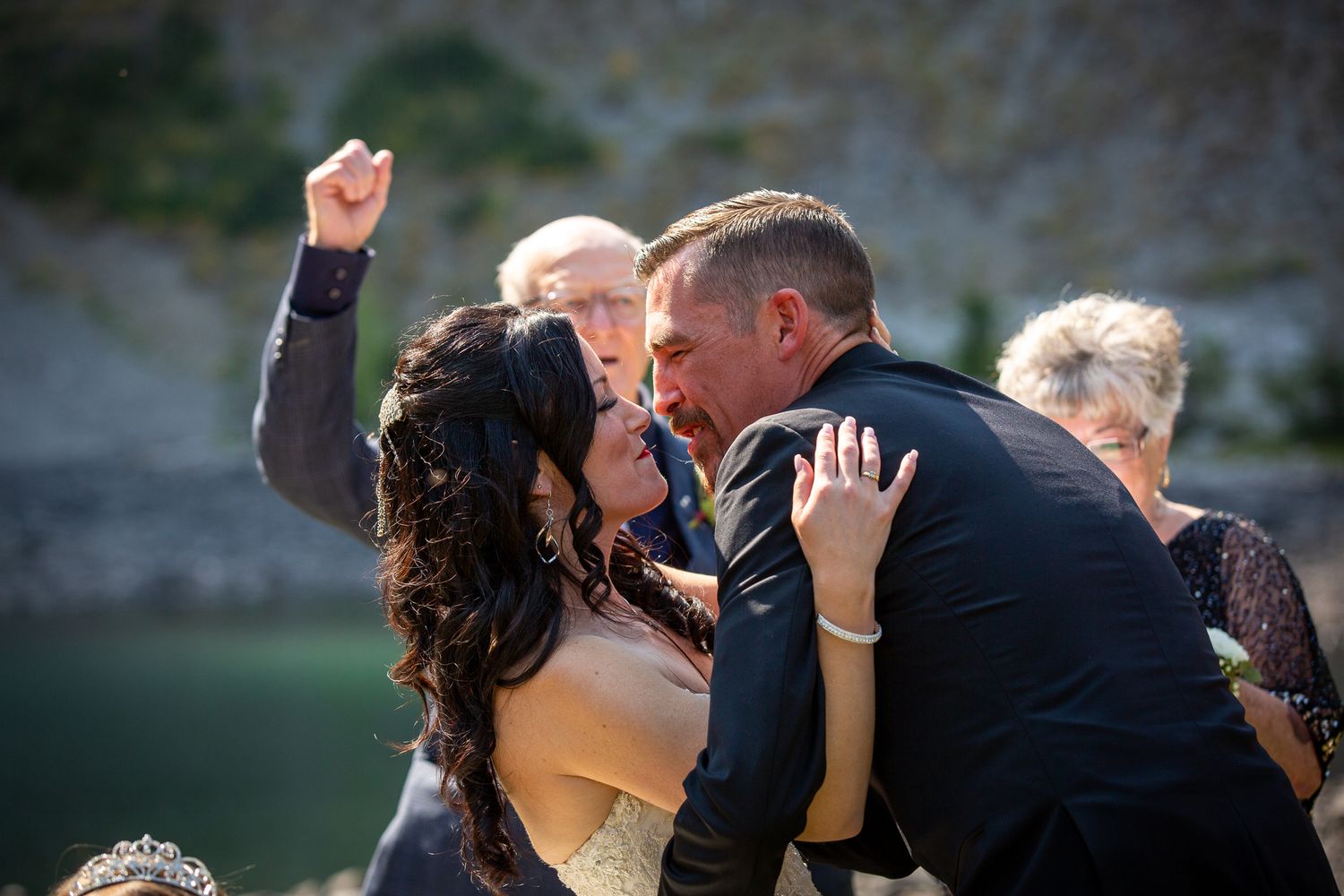 Bride and groom share a passionate kiss at outdoor wedding ceremony near a lake, with guests celebrating behind them in Kananaskis, Alberta.