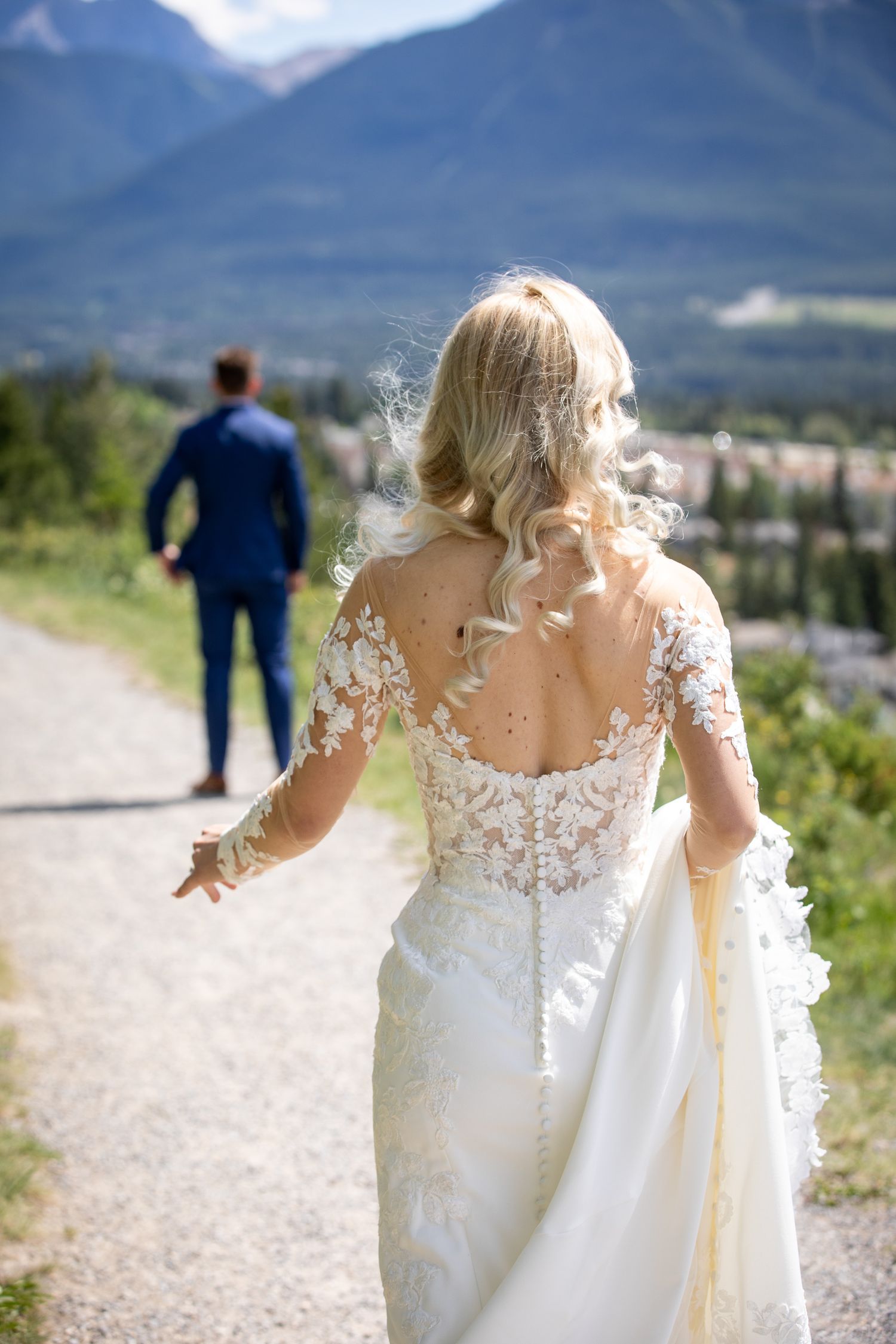 Bride in lace wedding dress walks toward groom in blue suit on mountain path with scenic valley backdrop In Canmore Benchland trail.
