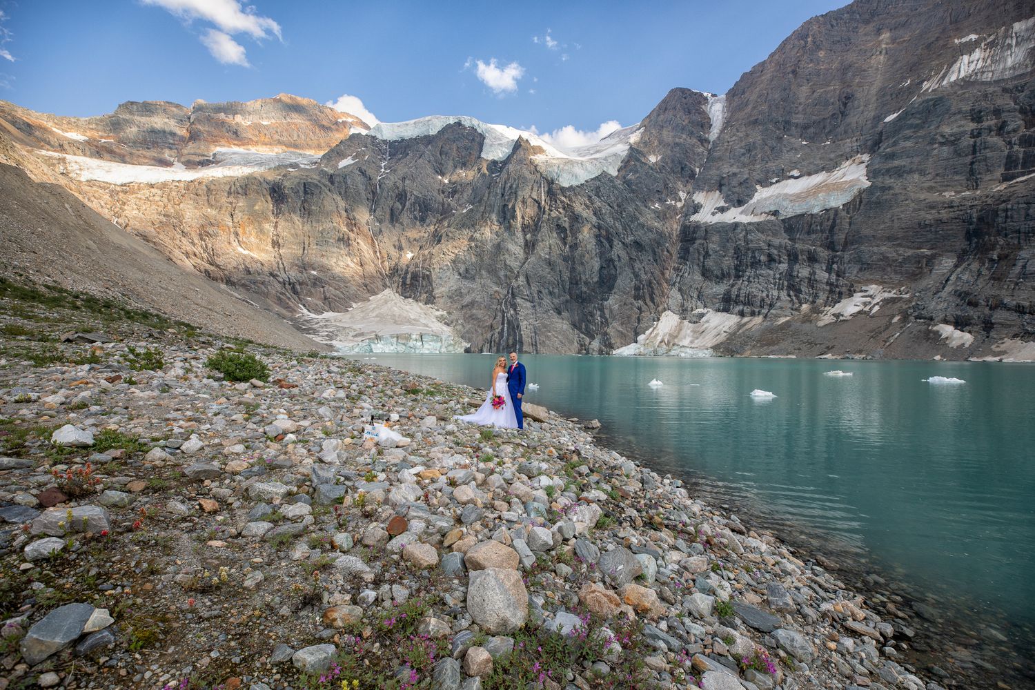 Couple eloping by a turquoise Lake-of-Hanging-Glacier surrounded by rocky mountain peaks and glaciers under a blue sky Panorama, BC.