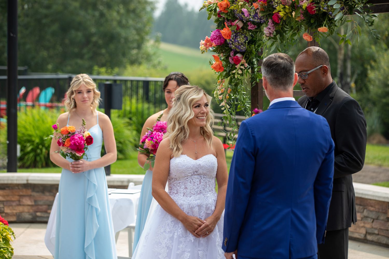 Outdoor wedding ceremony with bride in white gown, bridesmaid in blue dress, and officiant under colorful floral arch. Copperpoint resort,