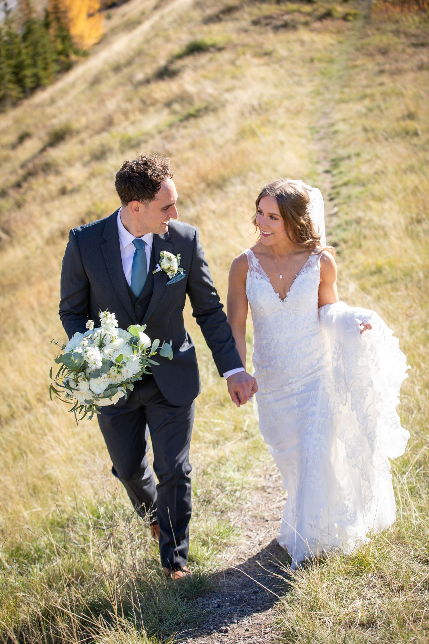 Newlywed couple walking through golden mountain meadow in formal wedding attire with white floral bouquet at Quarry lake in Canmore, AB.