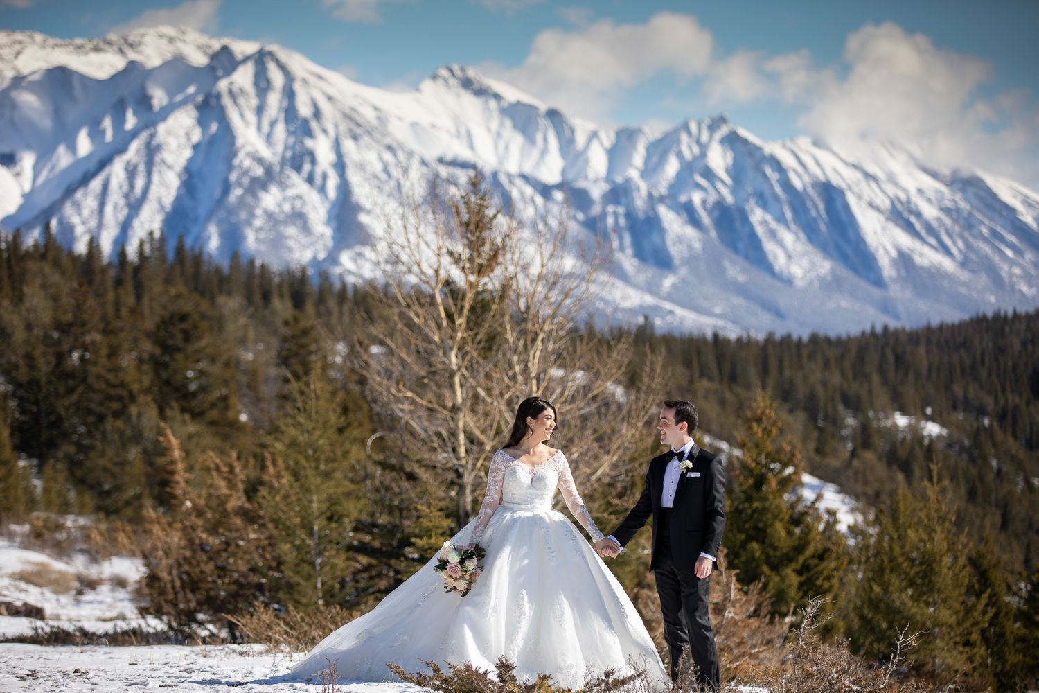 A couple in wedding attire poses against a snowy mountain landscape with evergreen trees and dramatic clouds at Tunnel reservoir in Banff.