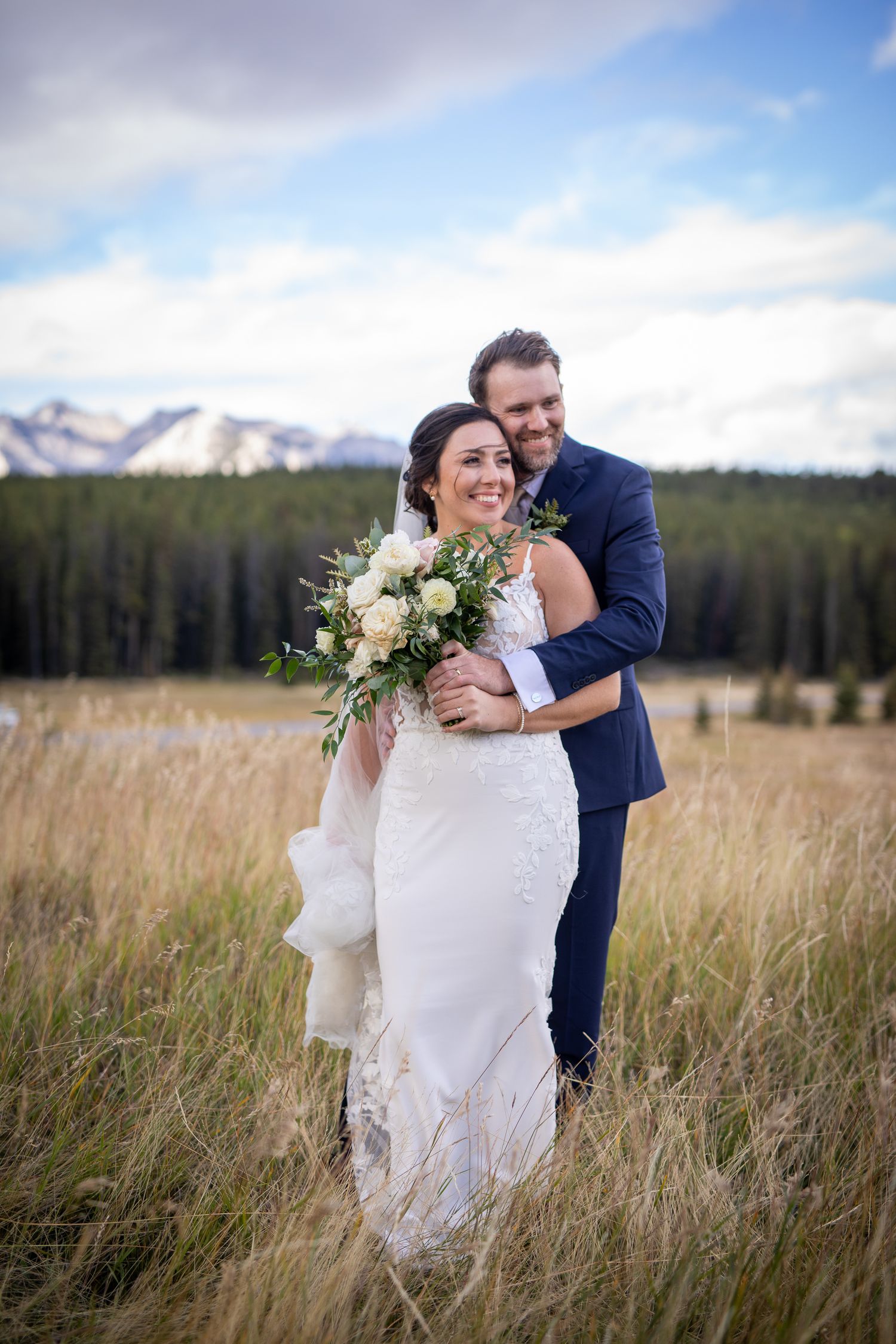 A couple embraces in a scenic mountain meadow with tall grass and evergreen forest on their wedding day. Banff, Alberta