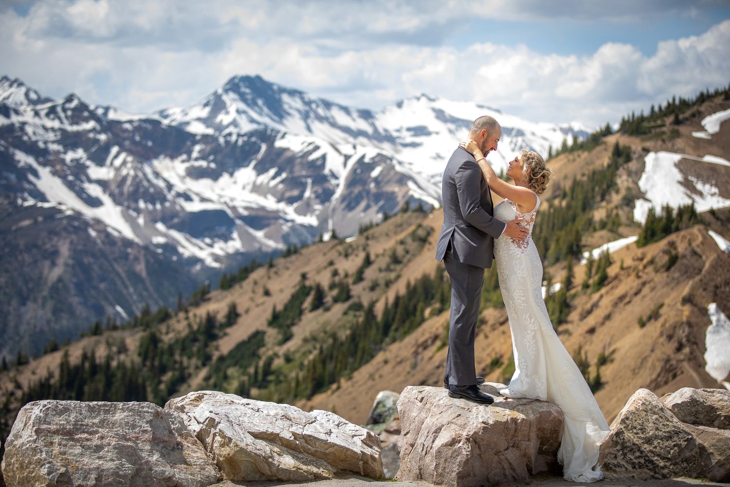 A couple shares an intimate moment on a mountain summit with snow-capped peaks in the background. Kicking Horse resort Golden, BC