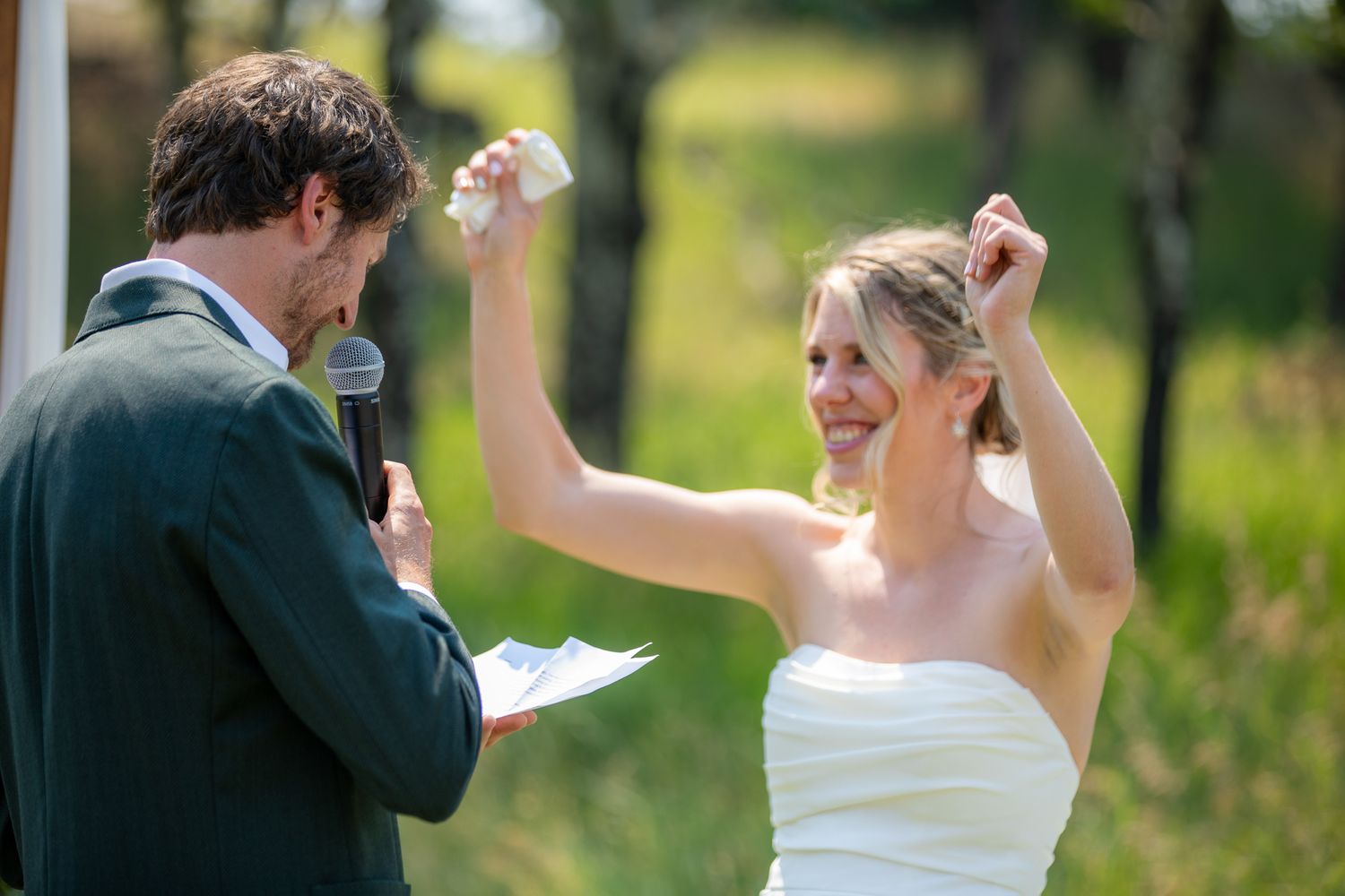 Joyful bride in white dress raises arms at outdoor wedding ceremony while groom reads vows into microphone at Swansea Ranch in Invermere, BC