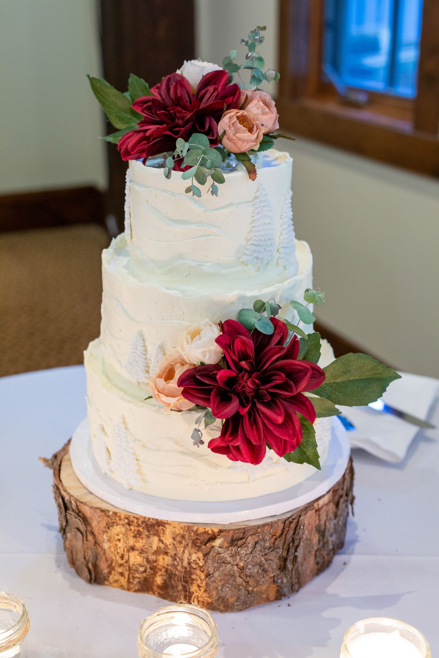 Three-tier white wedding cake with burgundy dahlias and greenery on rustic wood slice stand, surrounded by candles.