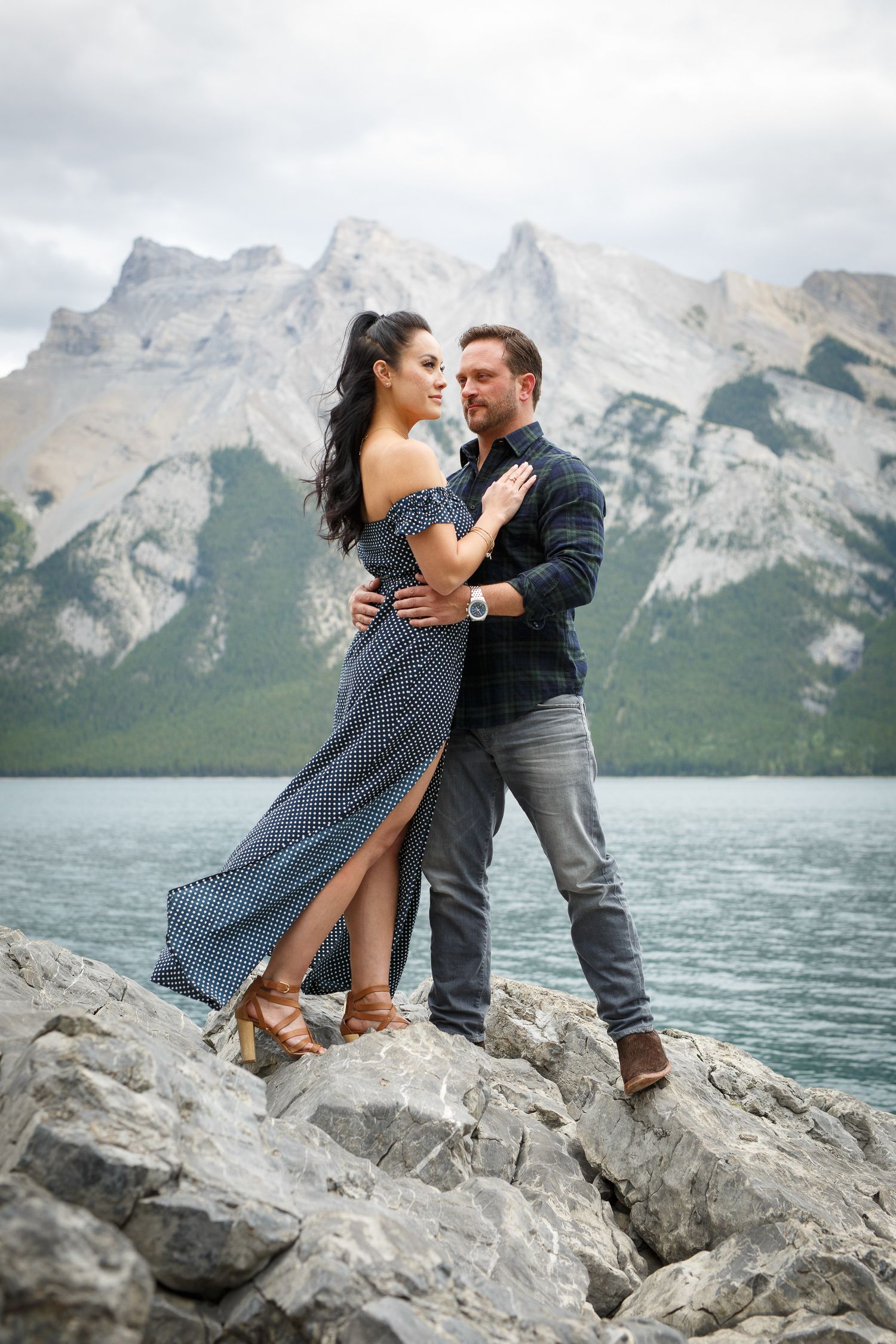 A couple embracing on a rocky shore with mountains in the background at Lake Minnewanka in Banff.
