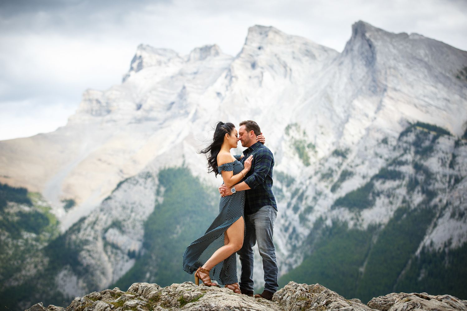 A couple embracing on a rocky outcrop with snow-capped mountains in the background at Lake Minnewanka in Banff.