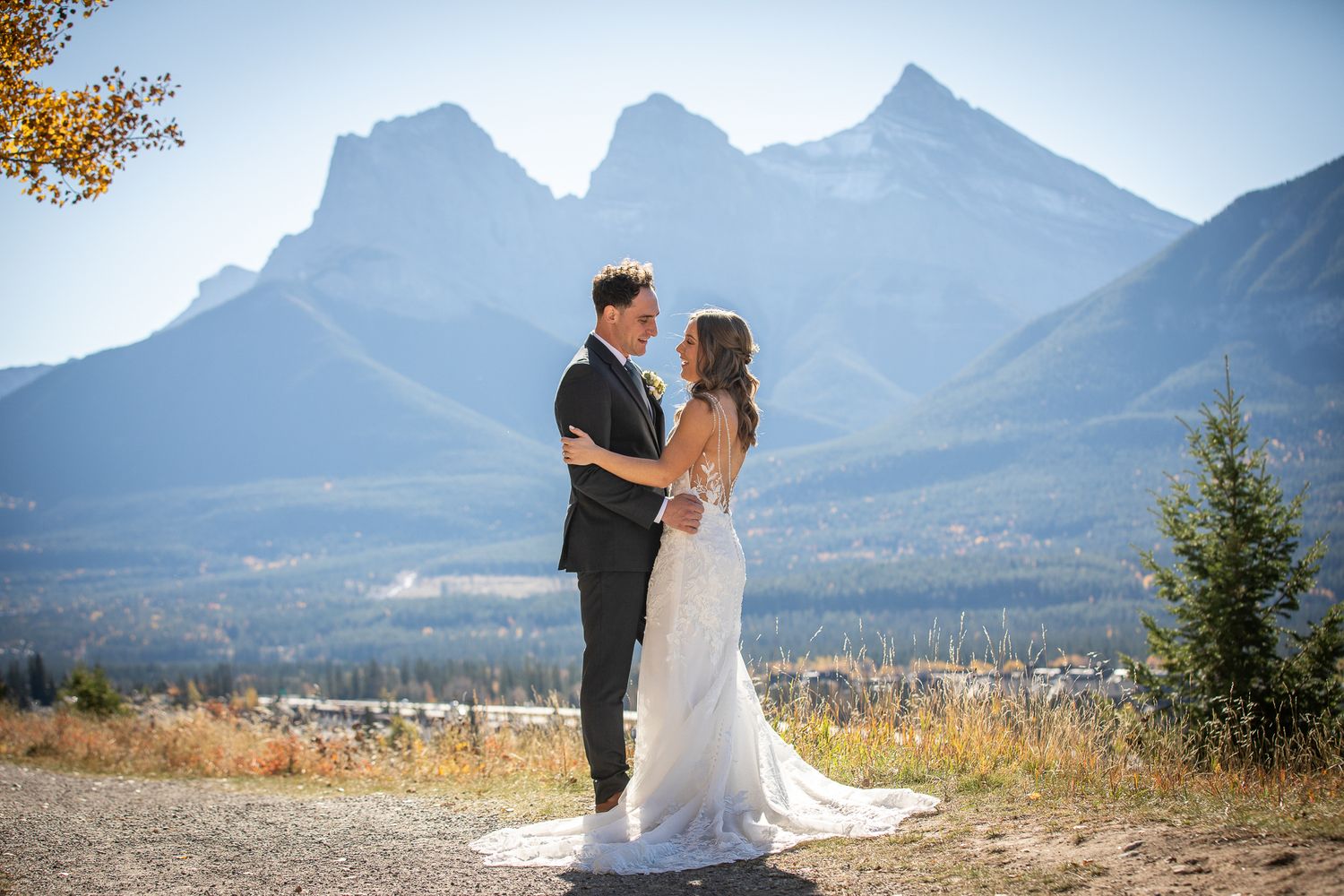 A couple in wedding attire embraces on a mountain overlook with Three Sisters peaks towering in the background in Canmore Benchland Trails