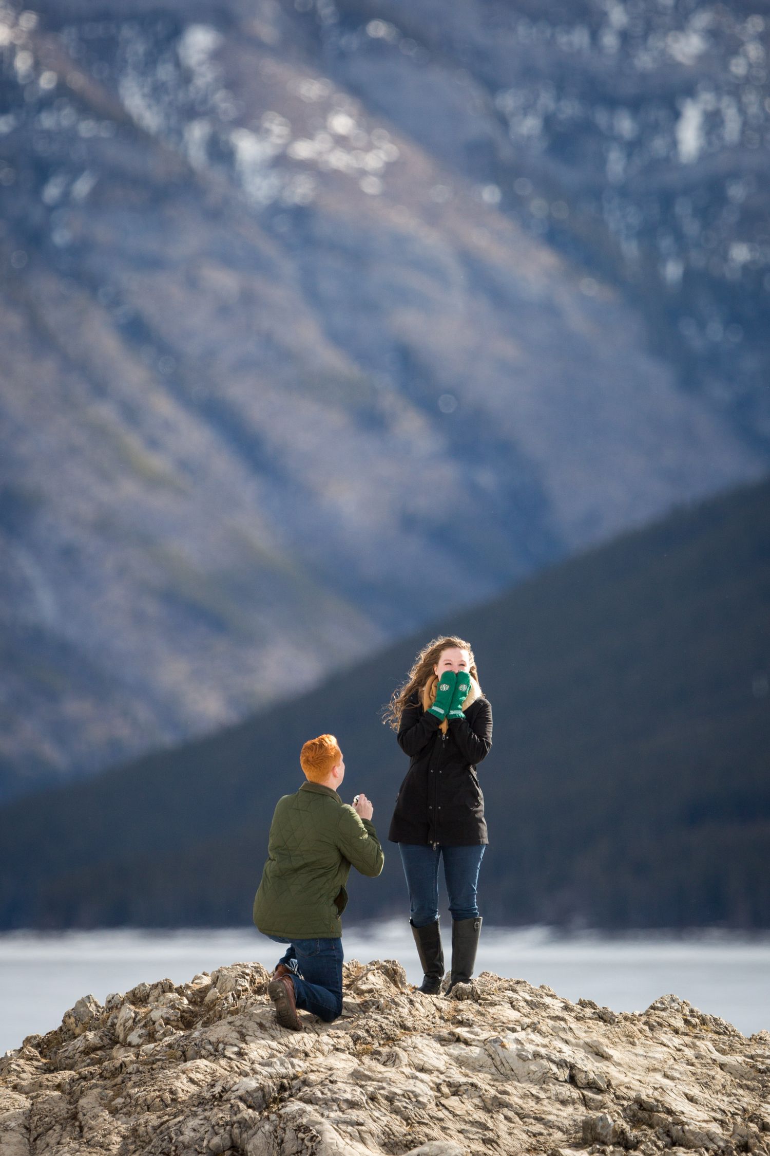 Man kneeling on rocky outcrop proposing to woman with mountains and lake in background at Lake Minnewanka in Banff National Park.