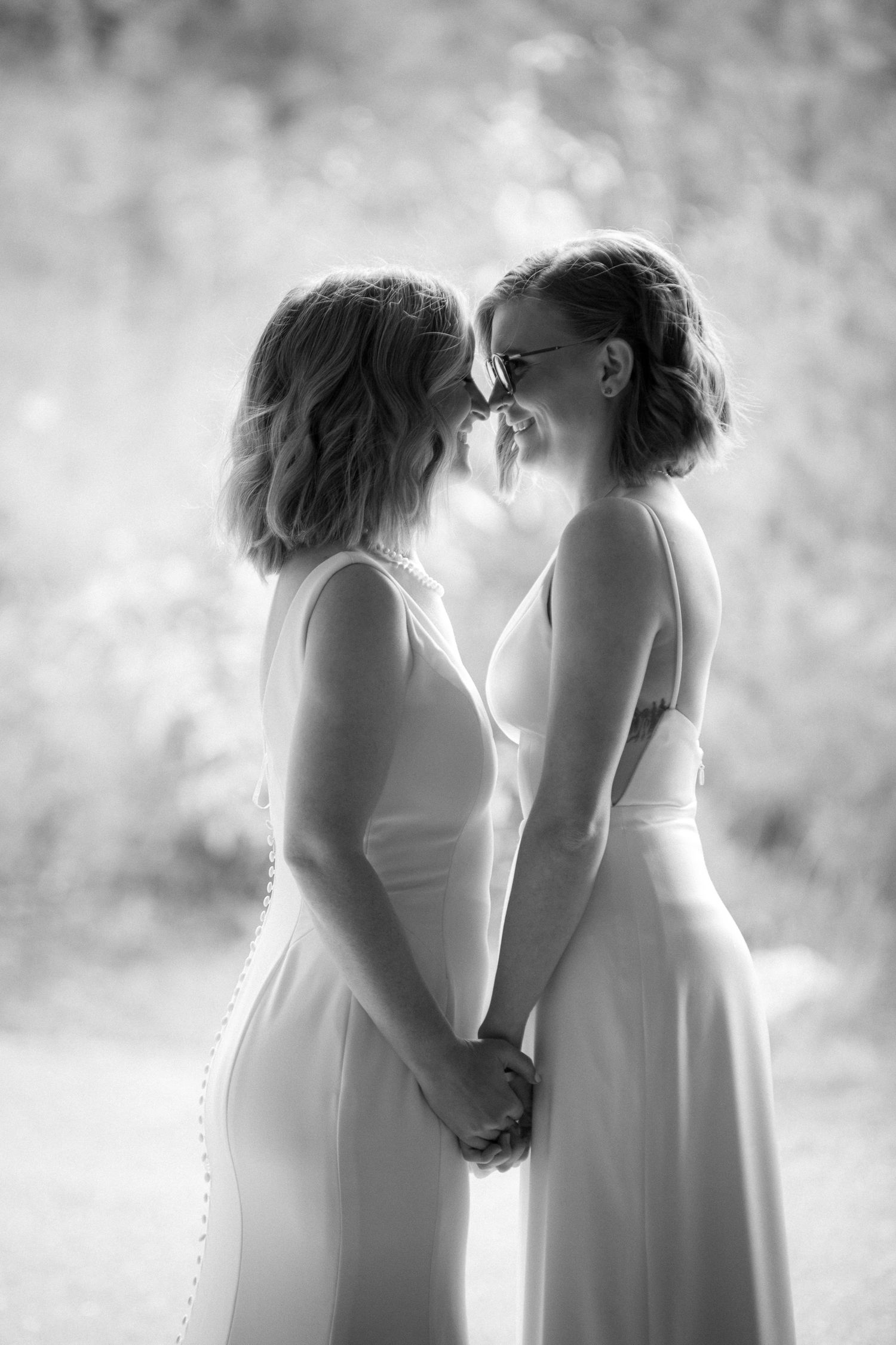 Two people in white dresses share an intimate moment in a dreamy black and white outdoor setting.