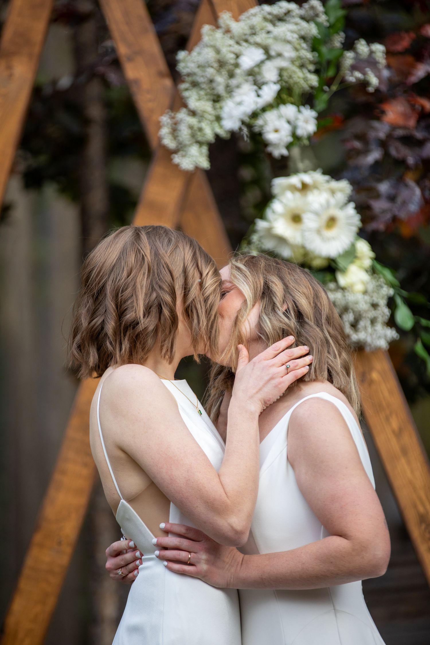 A LGBTQ couple in white dresses share an intimate moment in a garden setting with white flowers in the background. Earl Grey Lodge Panorama