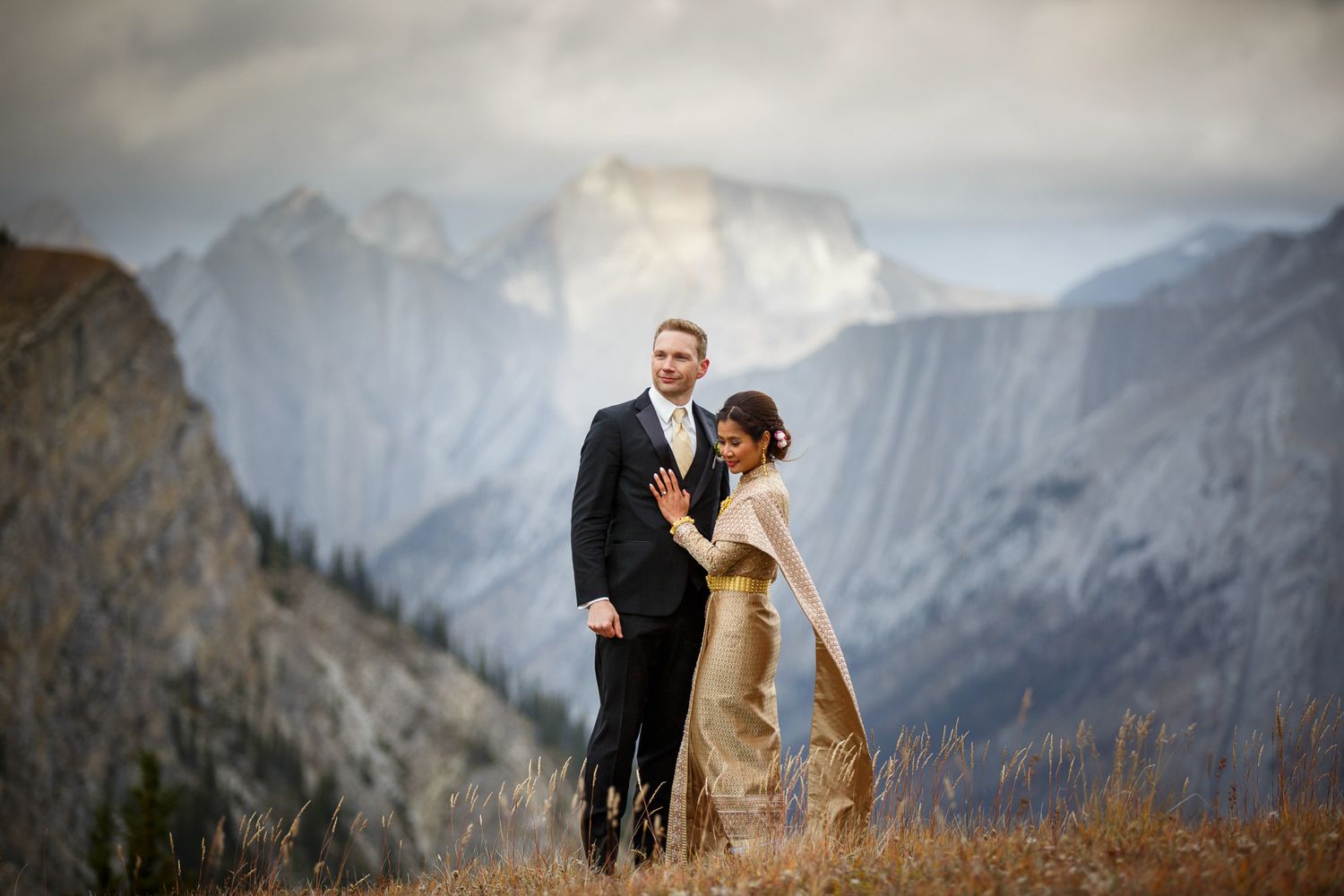 A wedding couple shares a romantic moment against the backdrop of majestic mountain peaks and dramatic cloudy skies in Banff, AB.