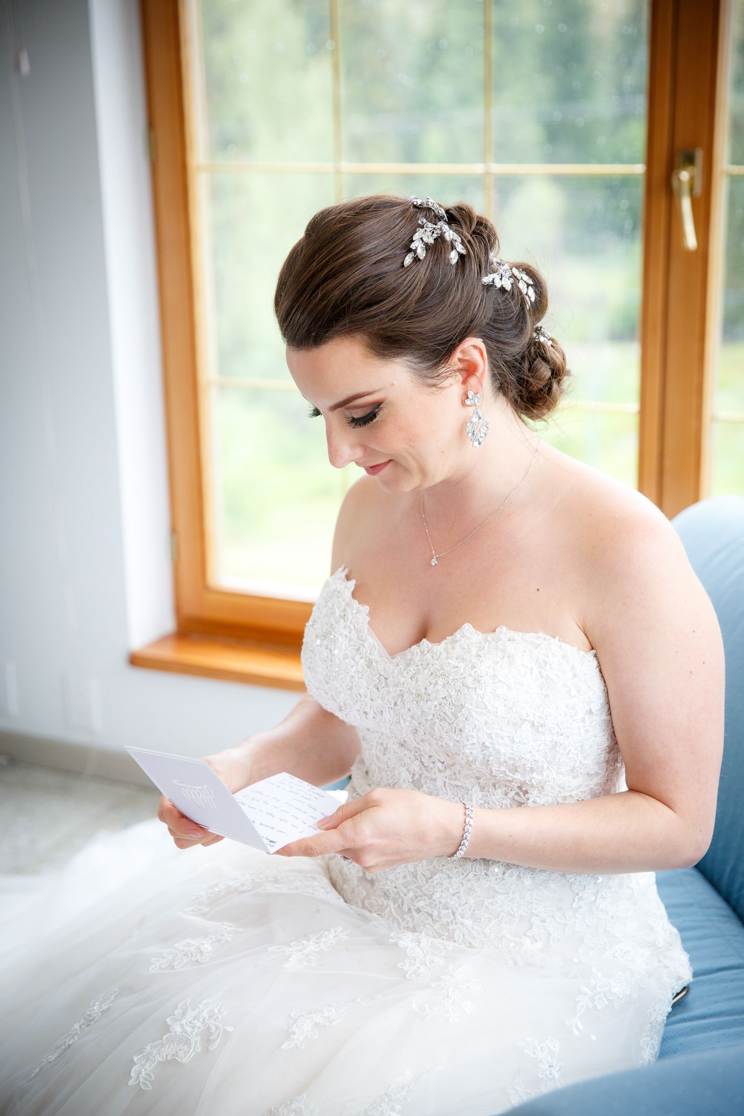 Bride in white lace strapless gown reads a letter, wearing elegant updo with hair accessories and chandelier earrings.