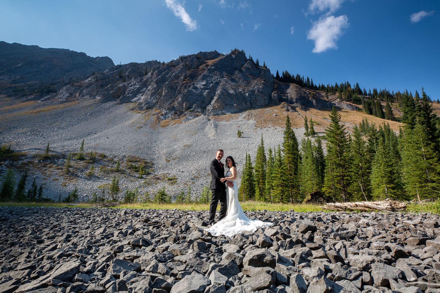 Bride and groom pose on rocky terrain with evergreen trees and mountain backdrop under blue skyin Kananaskis, Alberta.